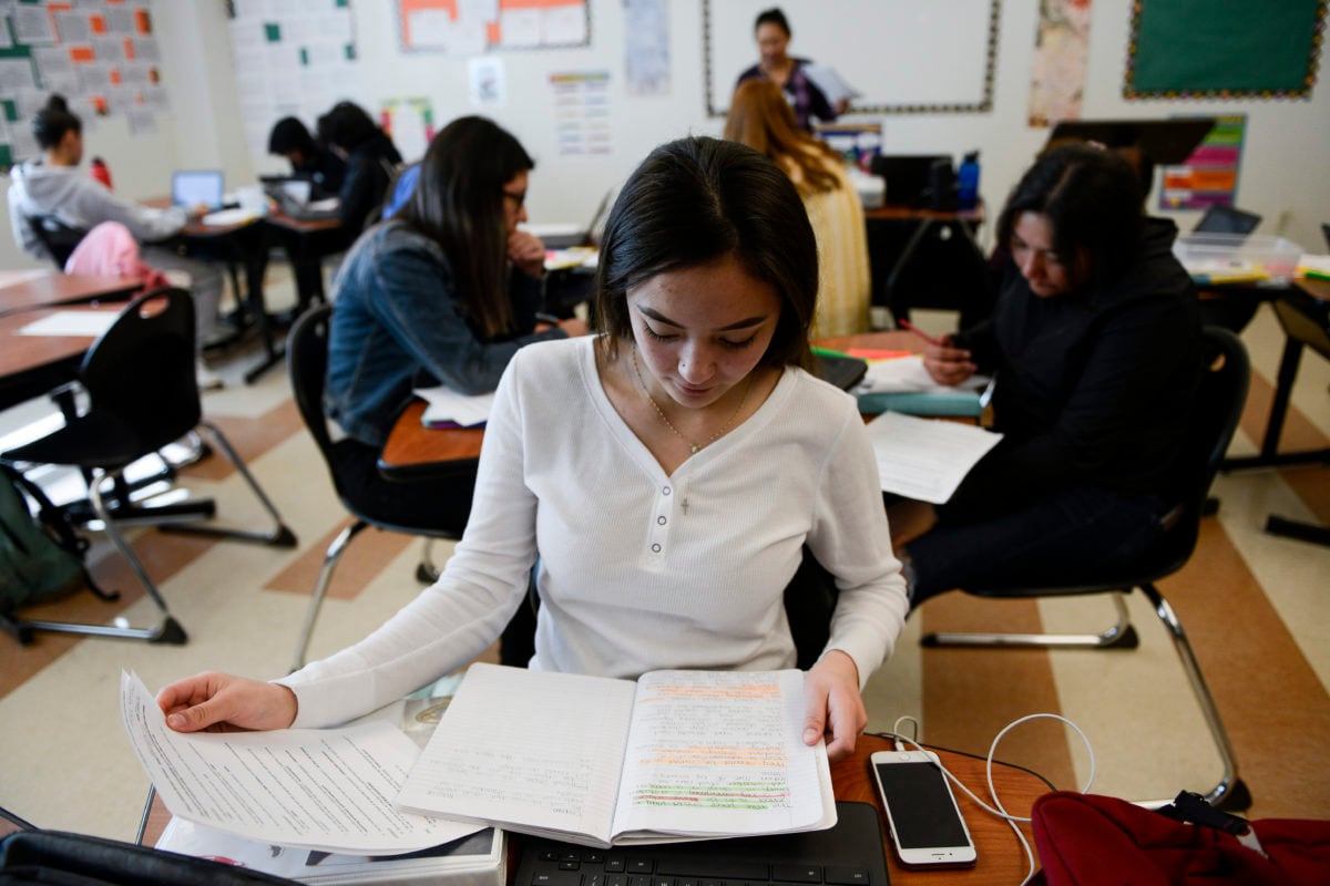 Adams City High School junior Alexandra Hernandez, in a white long-sleeve shirt, looks at her notebook, with a cell phone next to her. In the background are about five students working at tables and a teacher standing in front of a white board, in an AP language class on Feb. 4, 2019.