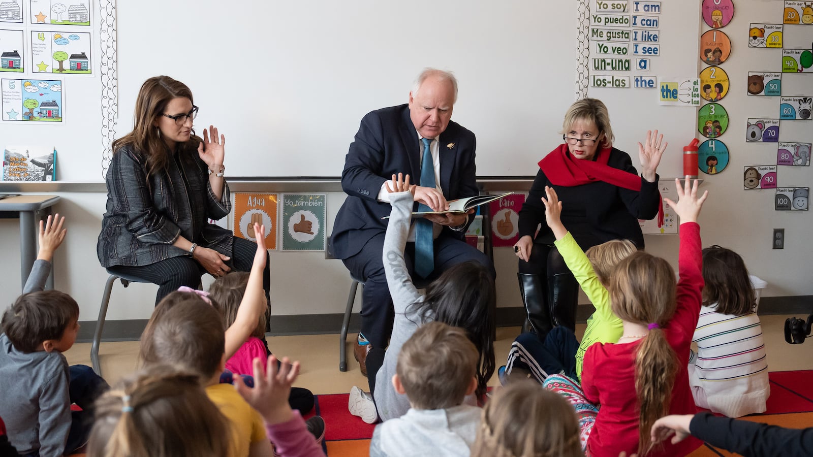 A man in a suit holds a book while sitting on a chair between two other adults and in front of a crowd of young students sitting on a colorful rug in a classroom.