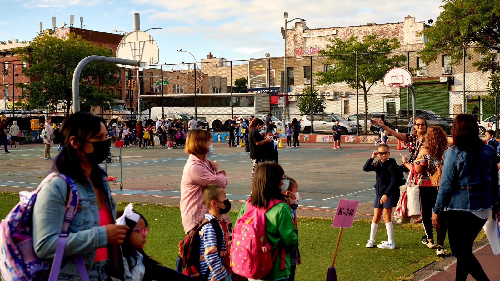 Parents and children walk outside of a school, with basketball courts visible in the background.