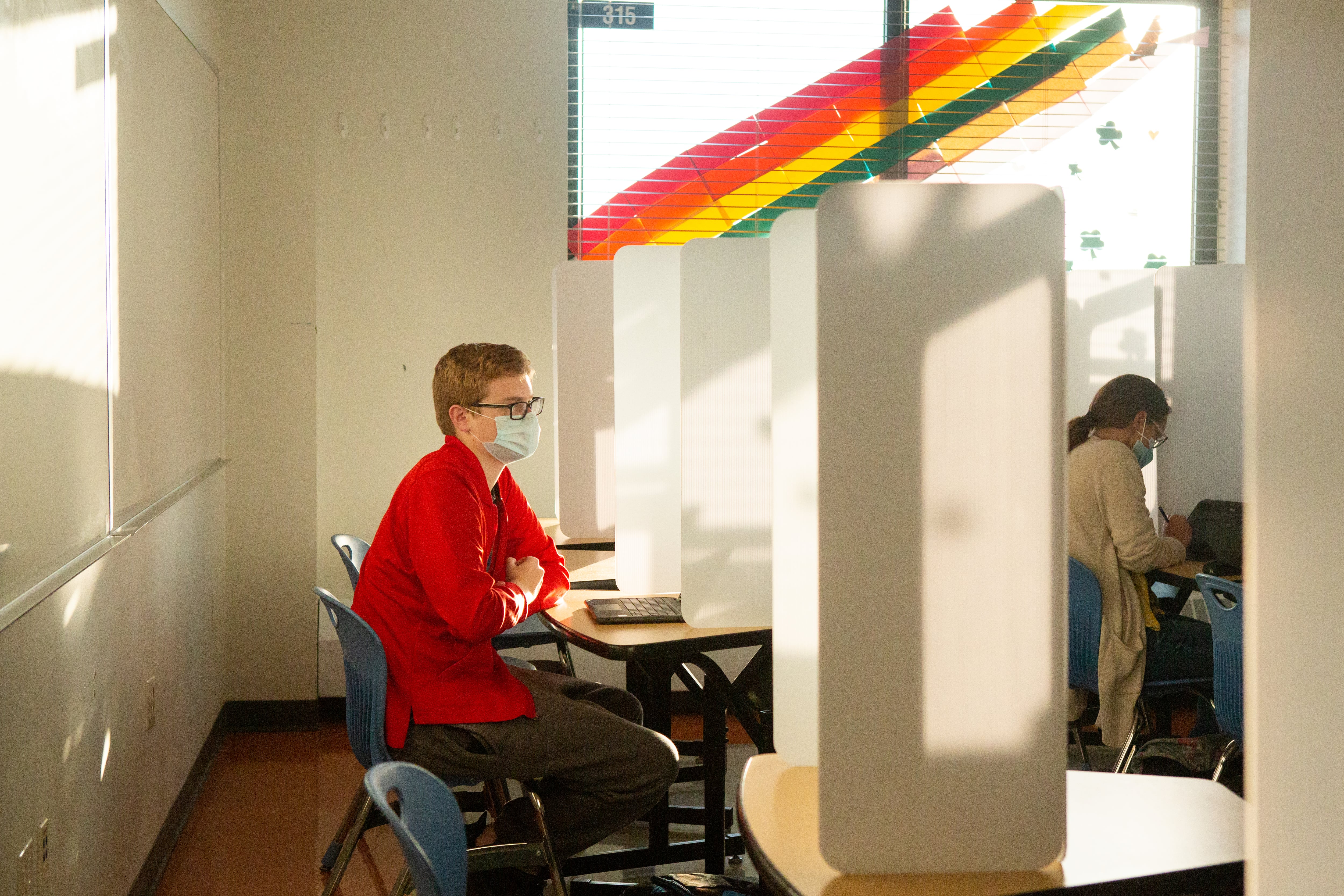 A masked teacher with glasses, wearing a red shirt and brown slacks, looks at a laptop as she sits at a desk separated by a folding white partition from the row in front where another person sits.