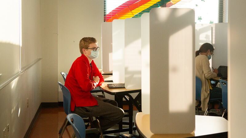 A masked teacher with glasses, wearing a red shirt and brown slacks, looks at a laptop as she sits at a desk separated by a folding white partition from the row in front where another person sits.