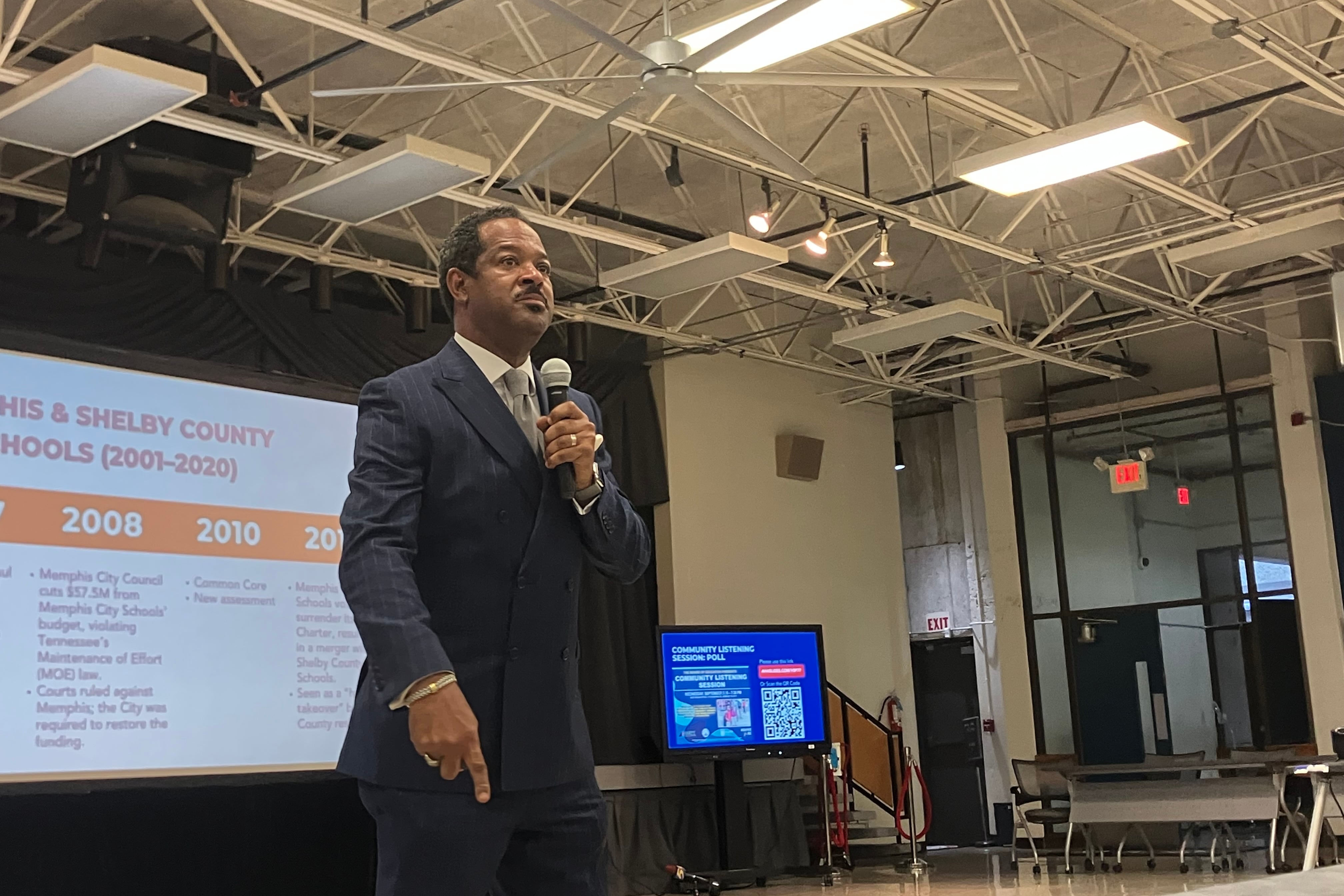 Superintendent Roderick Richmond, in a blue suit, stands with a microphone in the forefront of the photo. He is pointing down emphatically.