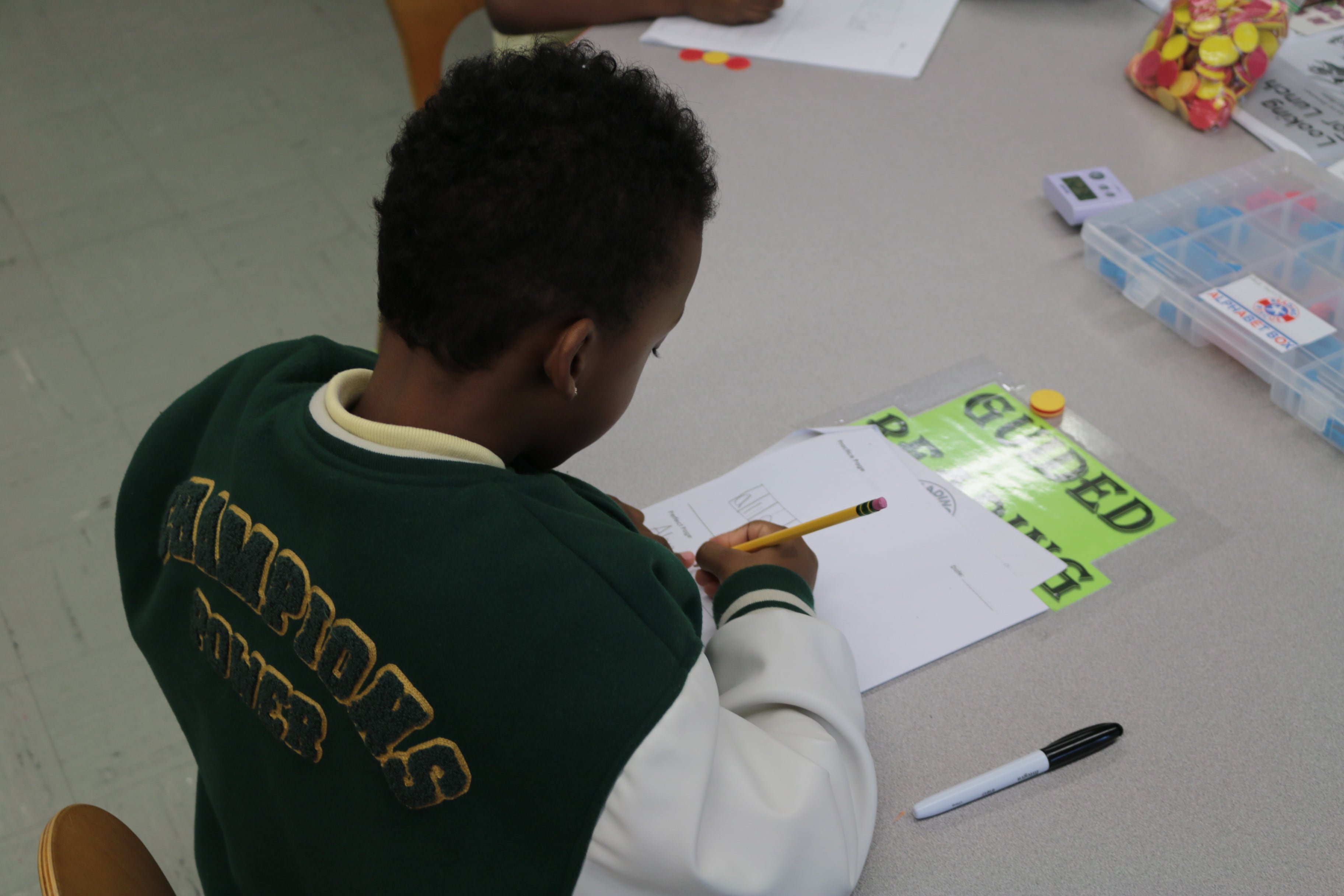 A young student wearing a dark green jacket sits at a table and works on a worksheet in a classroom.