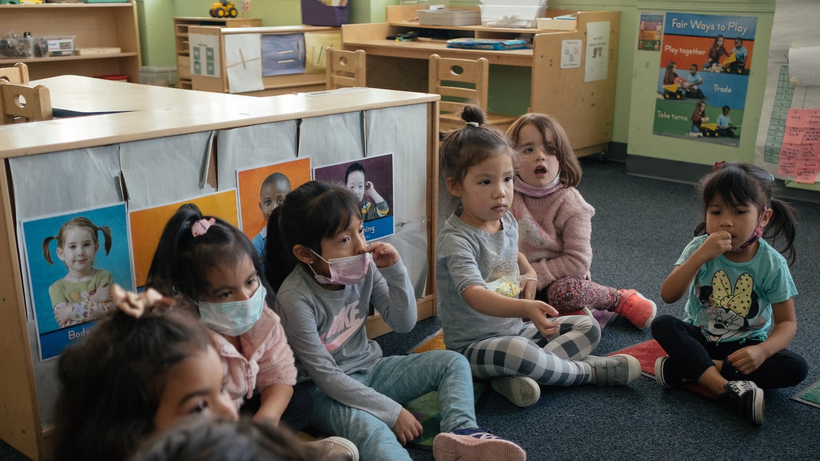 A group of students sit in a semi-circle on the floor of their classroom.