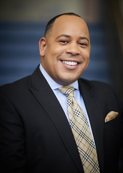 A man in a dark suit poses for a portrait in front of a blue wall.