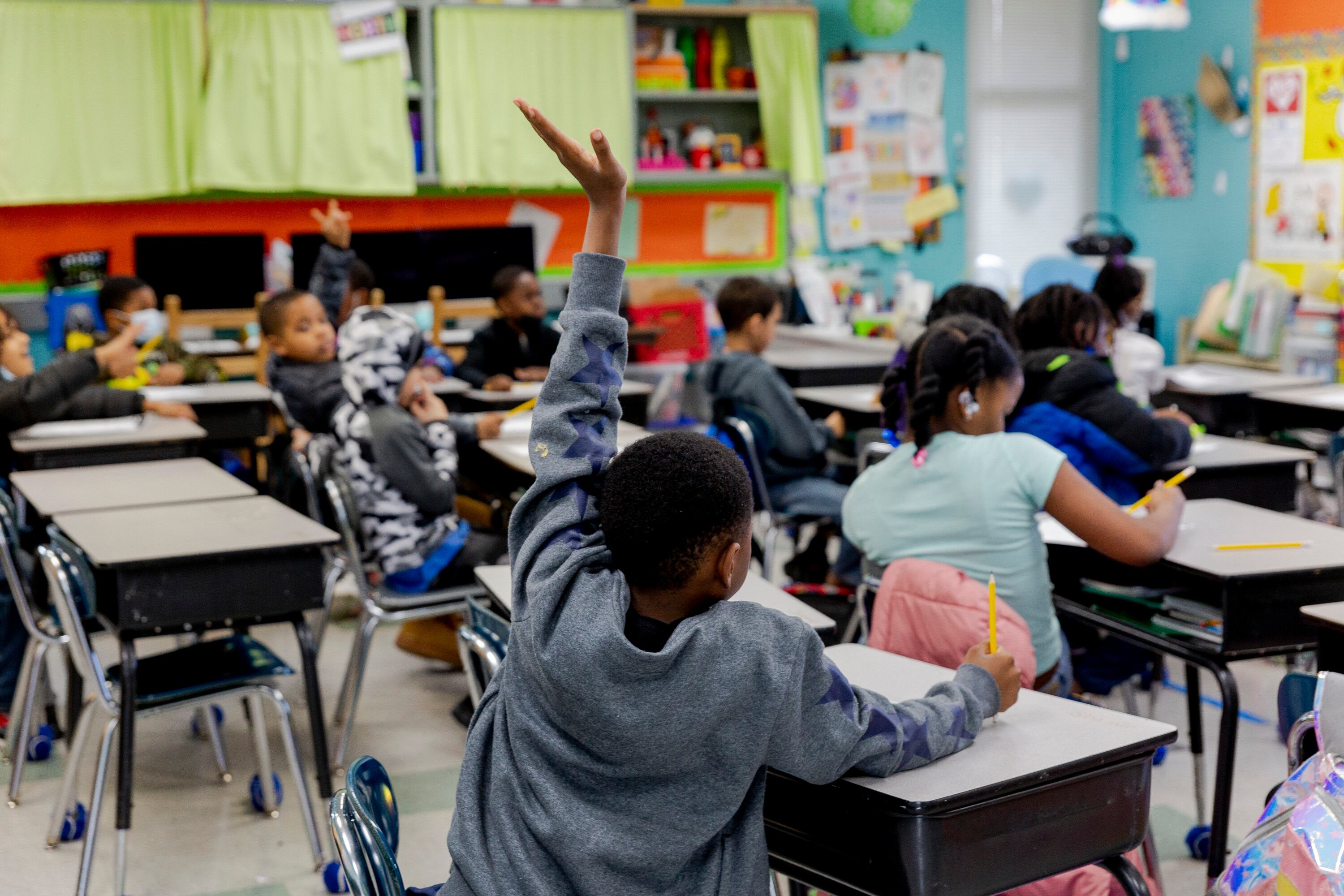A student sitting at a desk inside of a classroom full of students raises their hand.
