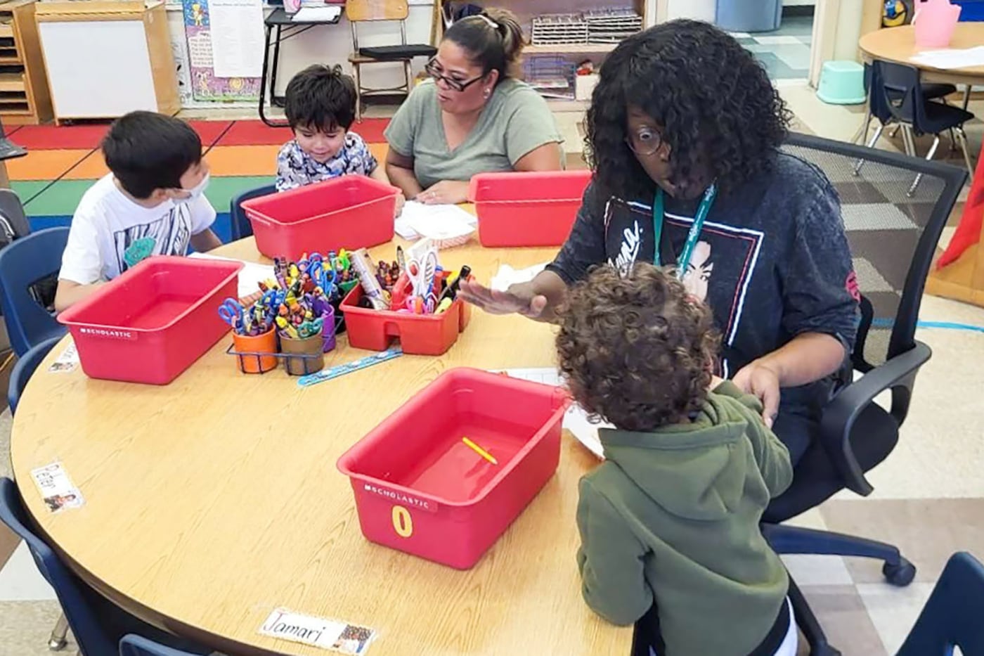 An adult teacher sits at a table with three young students in a classroom.