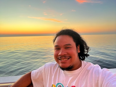 Headshot of a man wearing a white t-shirt. He appears to be on a boat just before sunset.
