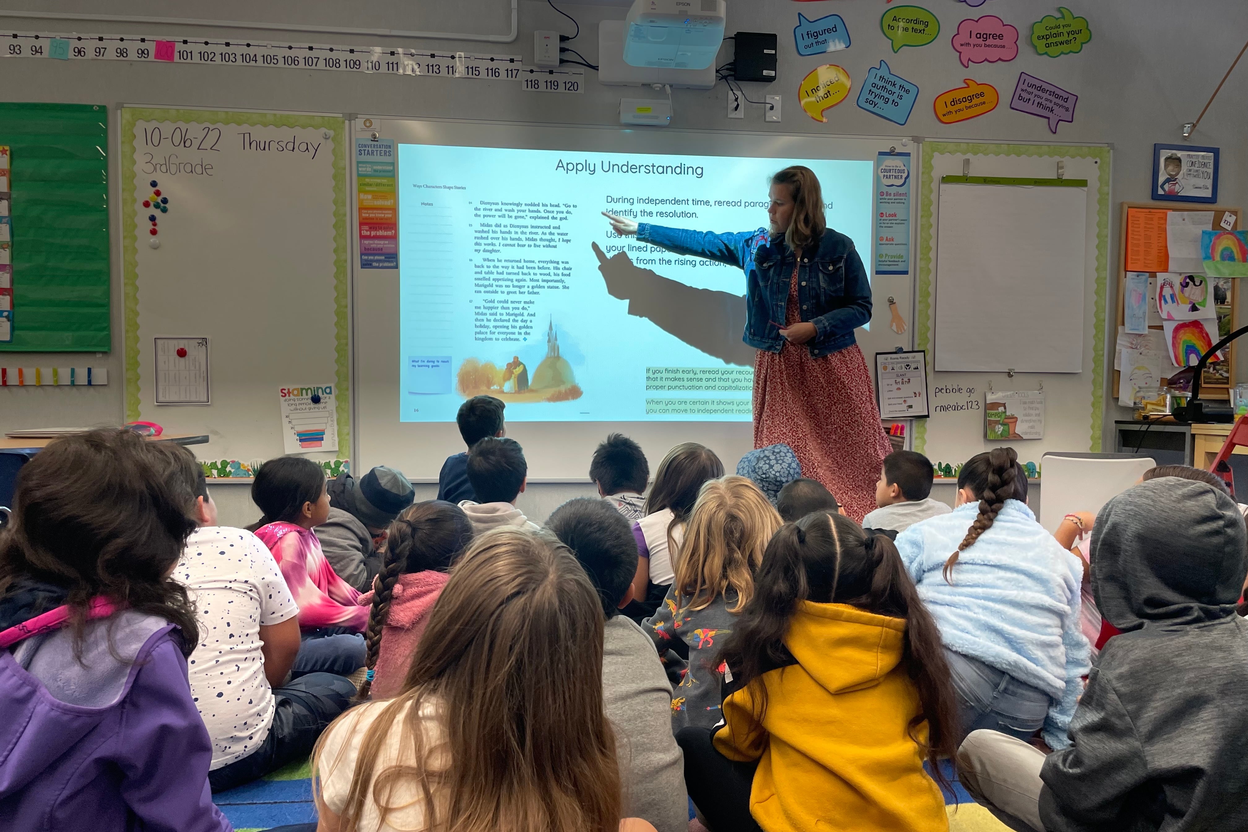 A teacher points to information on a white board as her students sit on the floor in front of her.