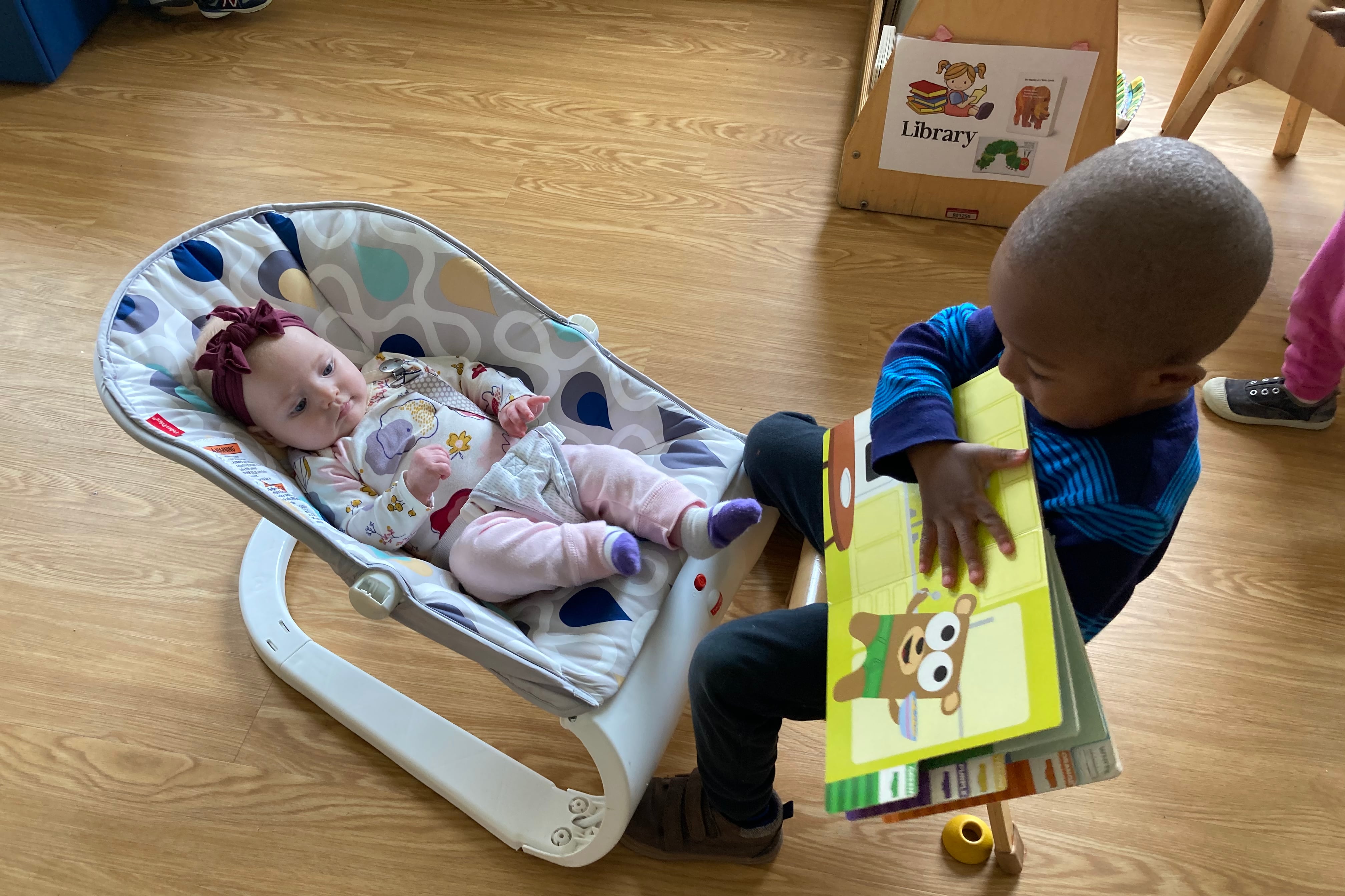 A young boy wearing a blue shirt reads to an infant wearing a purple bow and pink and white onesie in a small rocker.