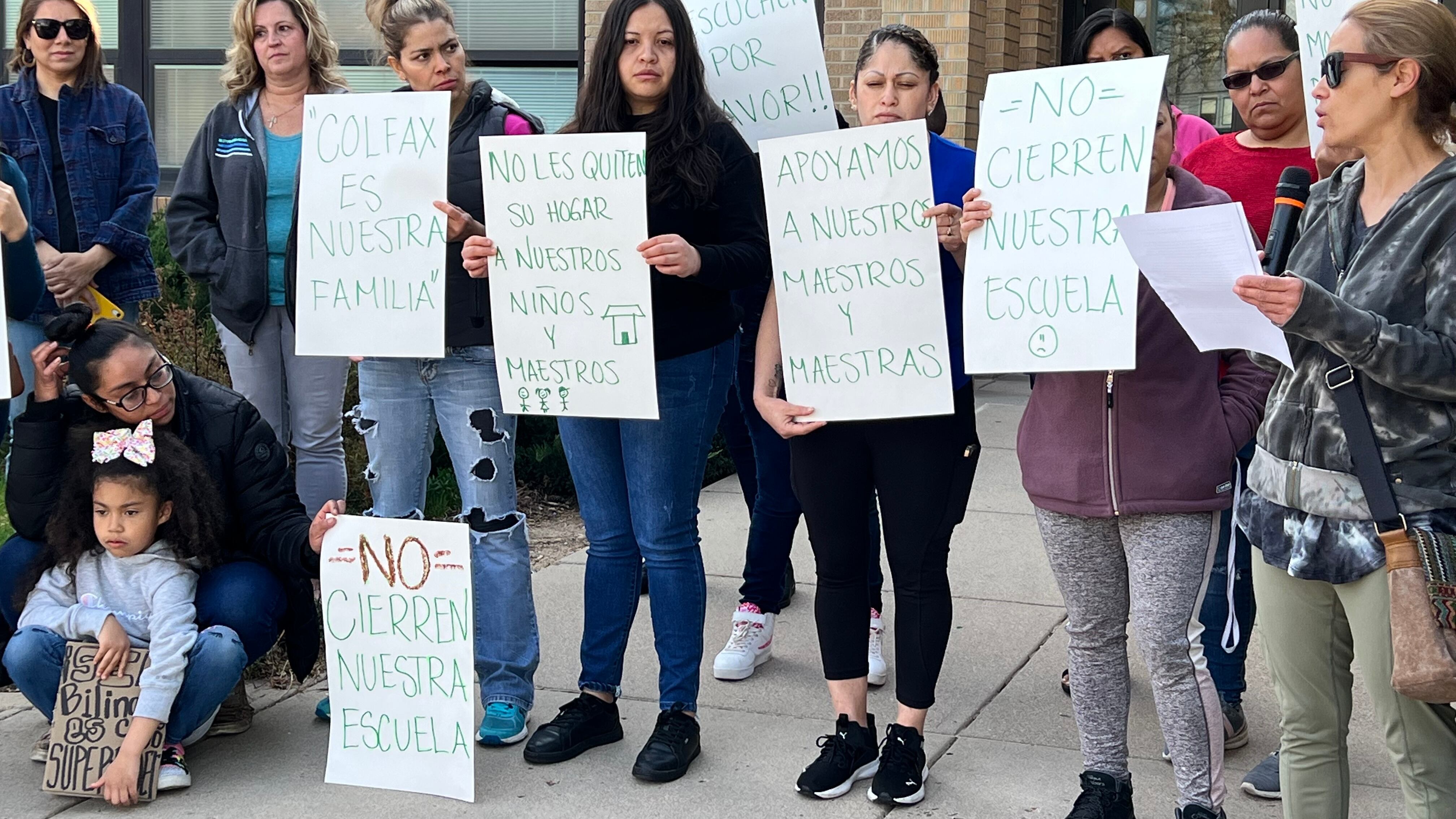 A group of women stand outside an elementary school. They hold signs that say “Don’t Close Our School,” “Colfax is Our Family,” and “DPS, Please Listen” in Spanish.
