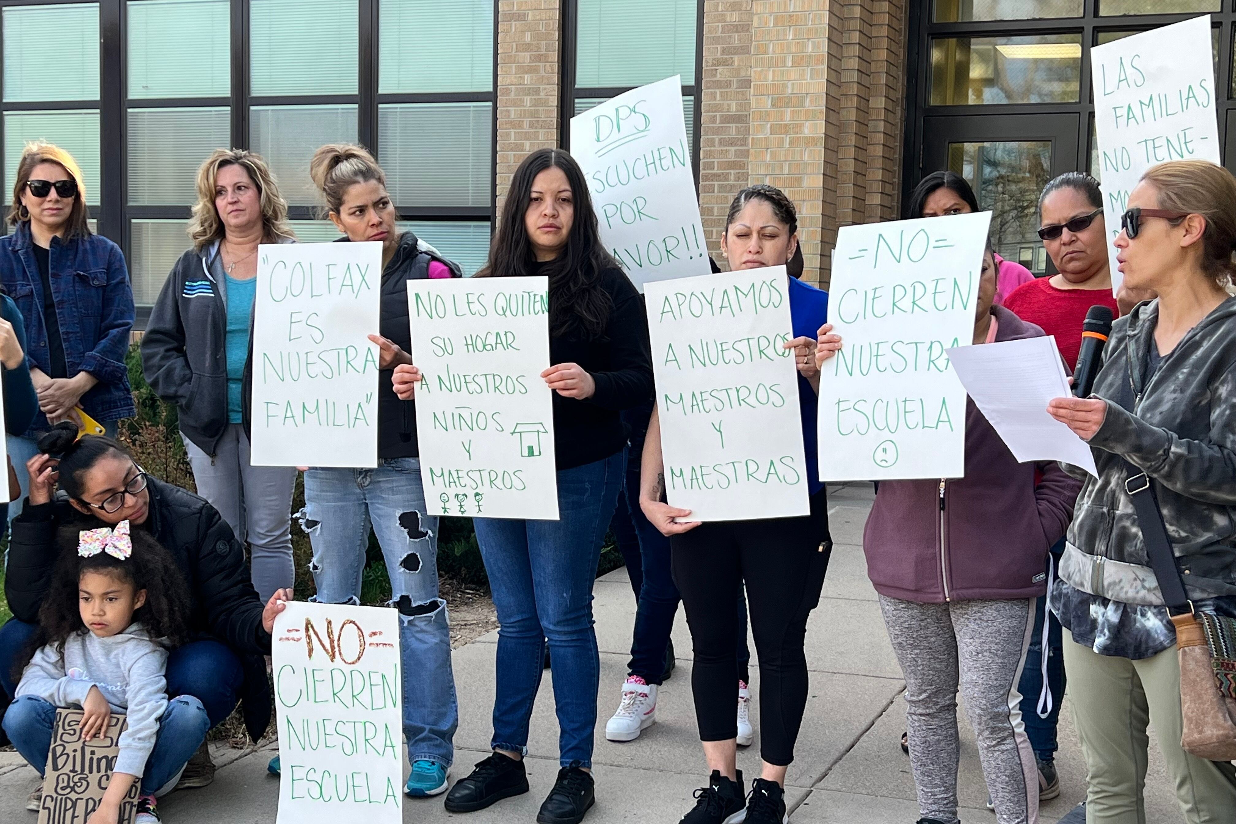 A group of women stand outside an elementary school. They hold signs that say “Don’t Close Our School,” “Colfax is Our Family,” and “DPS, Please Listen” in Spanish.