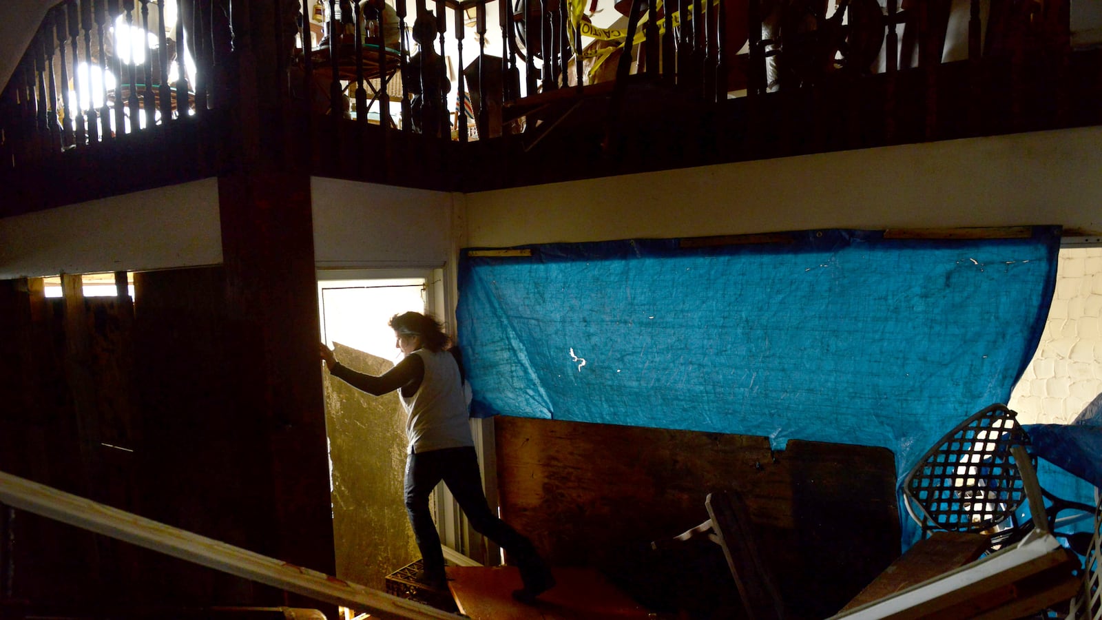 A woman stands in the doorway of a home with blue tarp hung on the wall next to her and fallen wooden beams in the foreground.