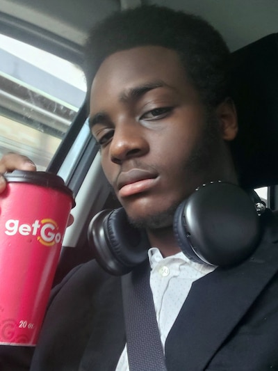 A Black high school senior takes a selfie in a car while holding a red to go coffee cup and headphones around his neck.