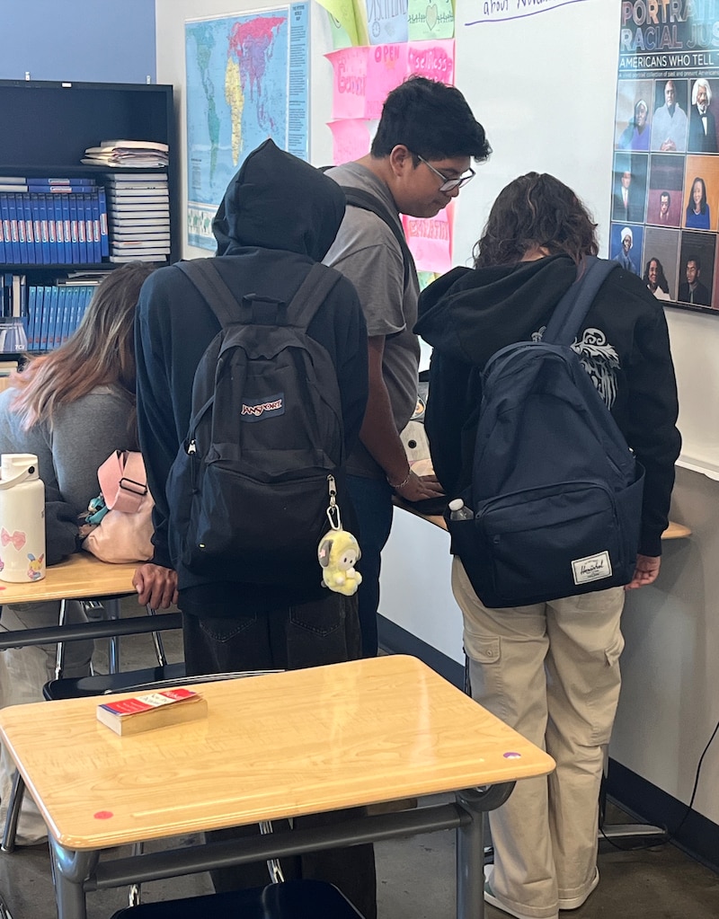A group of high school students wearing backpacks gather around a book filled with old CDs.