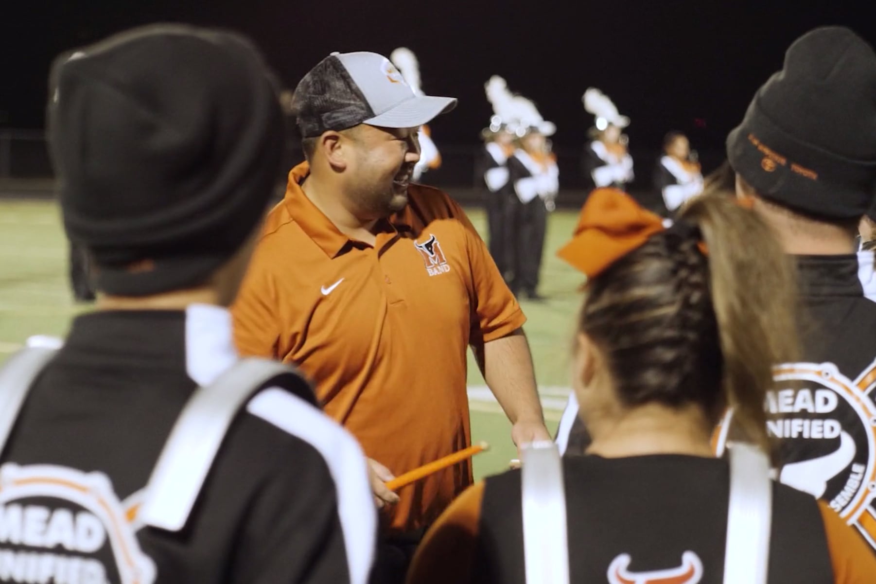 A teacher in an orange shirt talks with students wearing marching band drums outside.