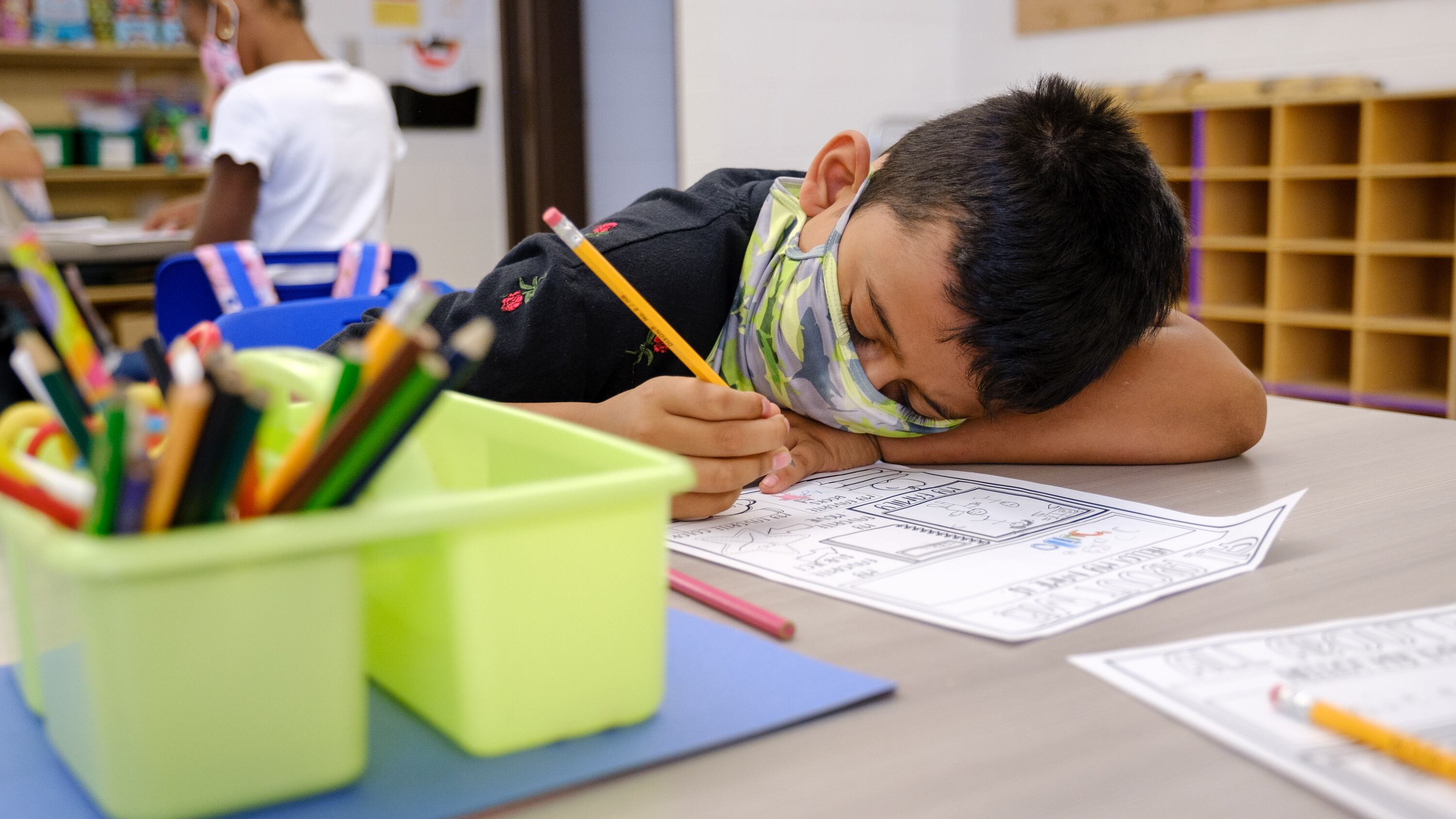 A young boy wearing a mask rests his head on a desk as he completes a worksheet.