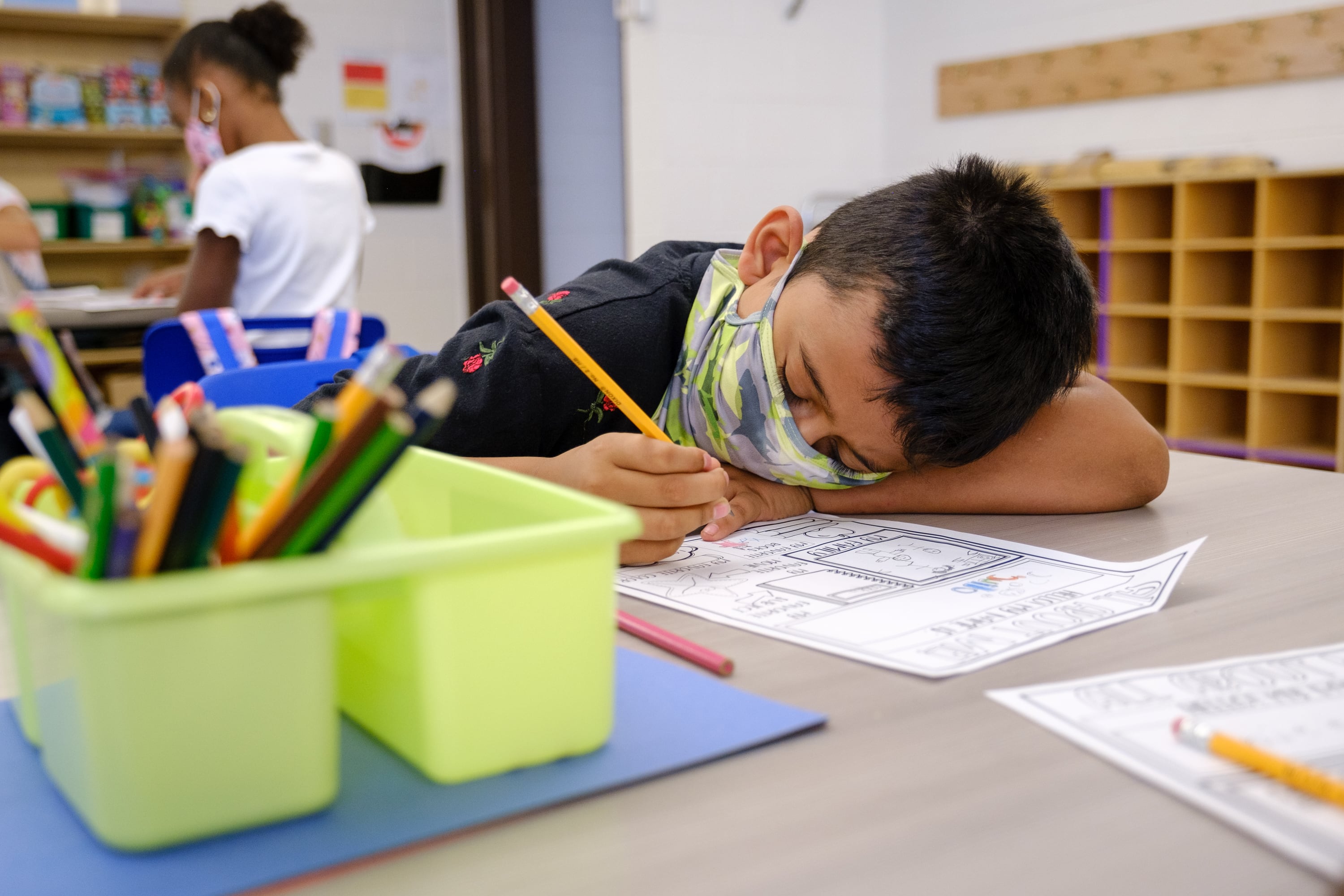A young boy wearing a mask rests his head on a desk as he completes a worksheet.