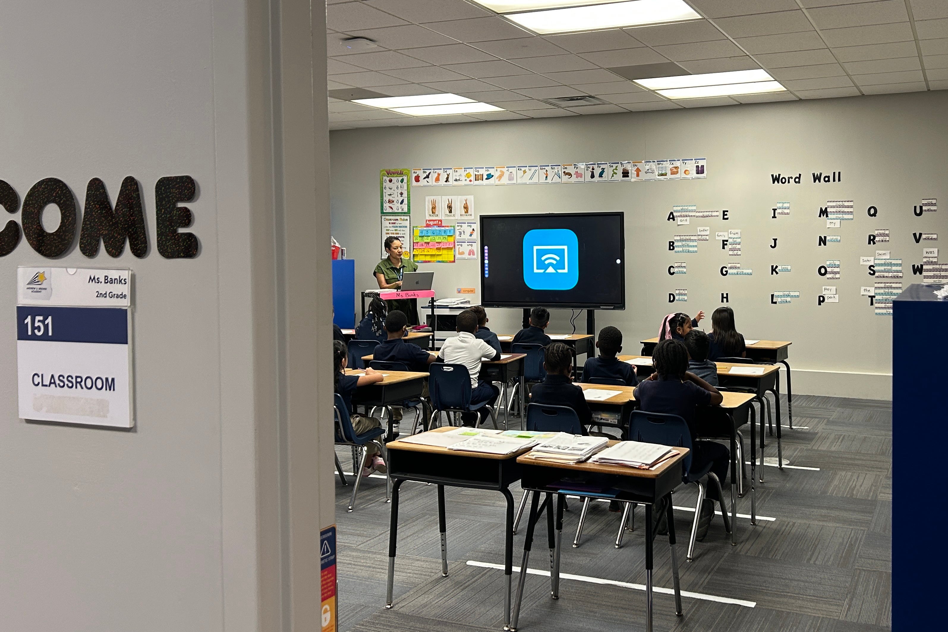 A photograph of a classroom with young students sitting in chairs at desks.