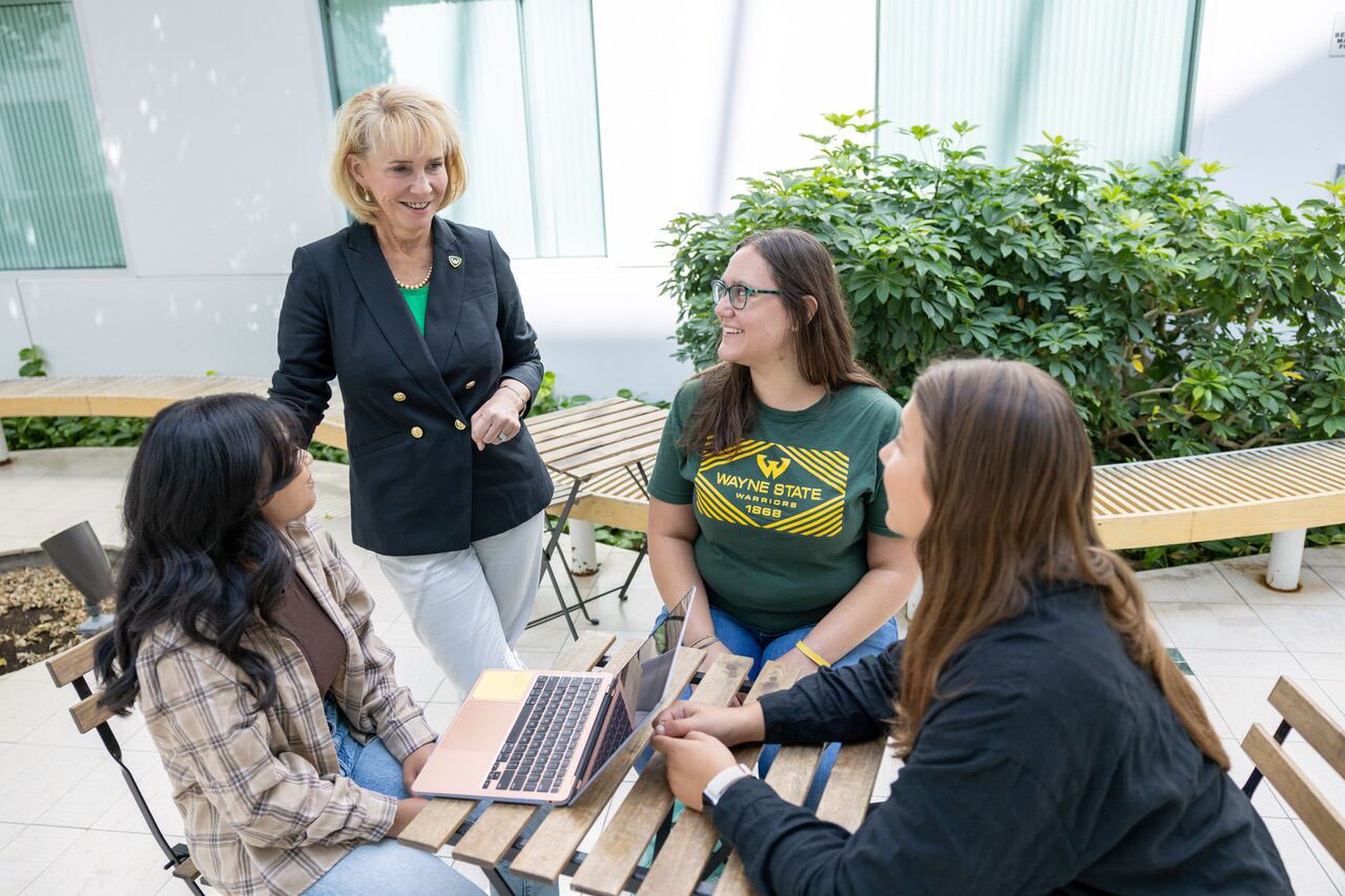 A blonde woman wearing a dark colored suit jacket, white pants and a turquoise top is standing up, talking to three students seated at a table.