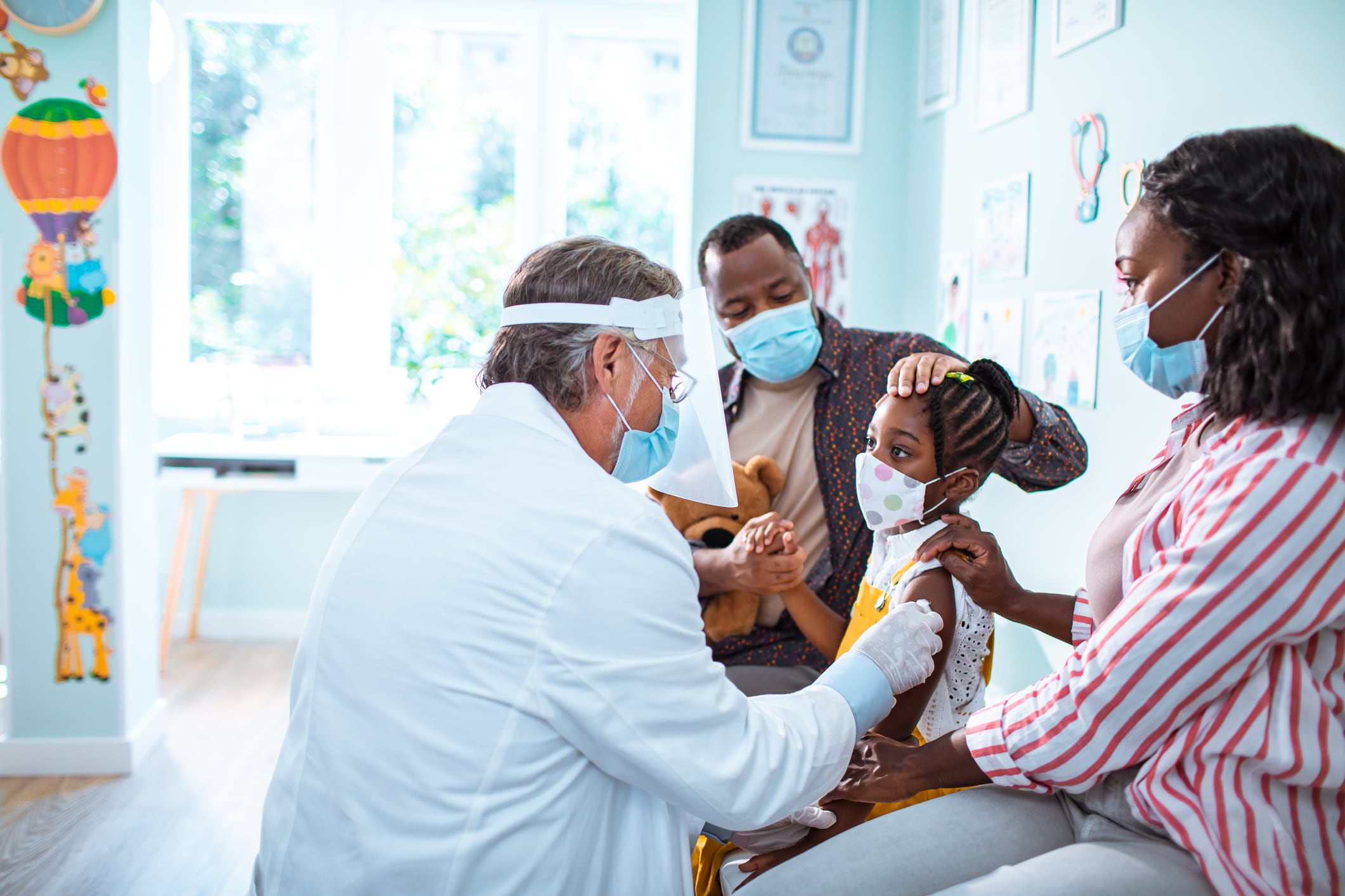A child receives a vaccine from a pediatrician wearing a white coat and face shield. Her parents sit on either side of her.