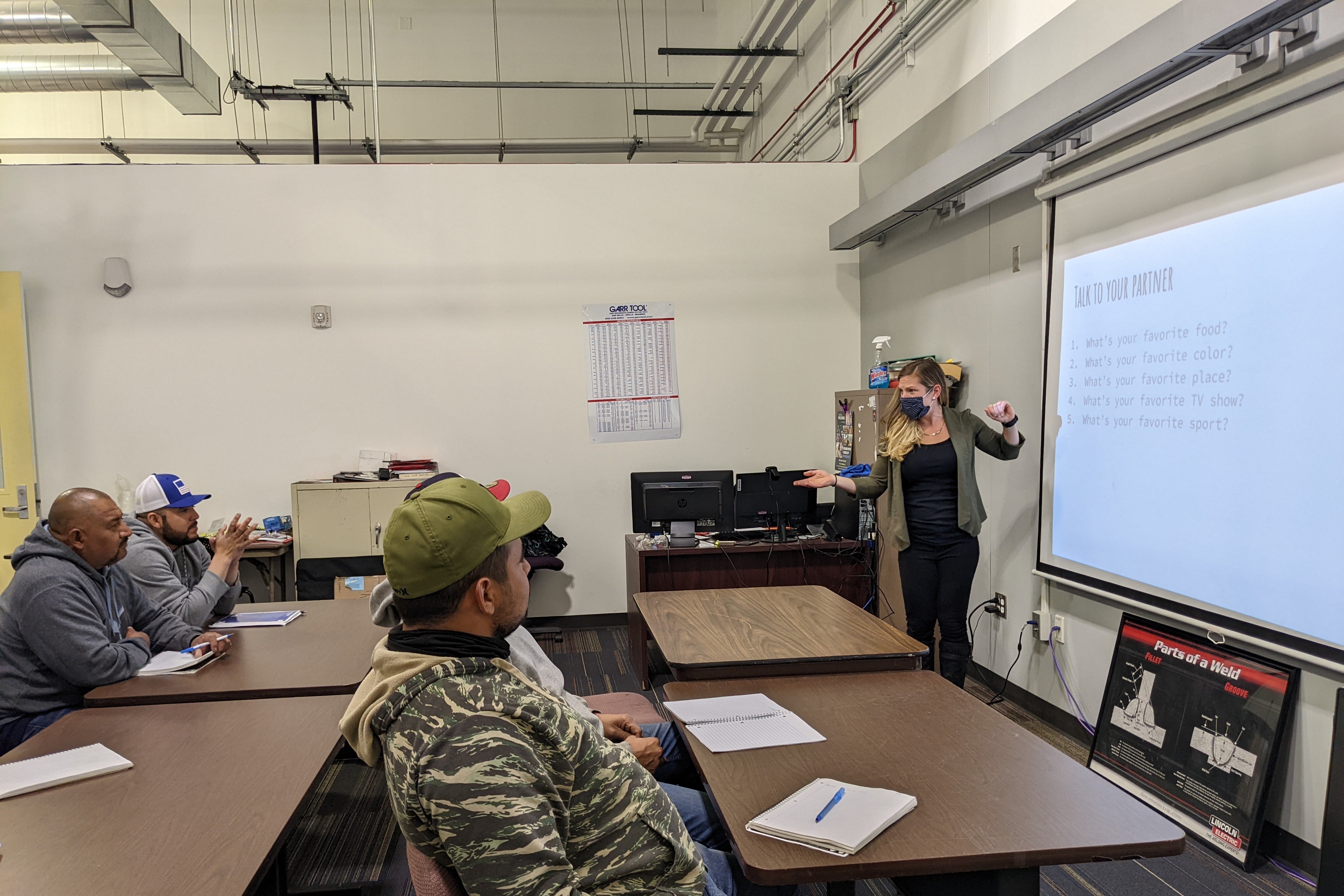 A woman stands in front of a white marker board and a group of students in a classroom.