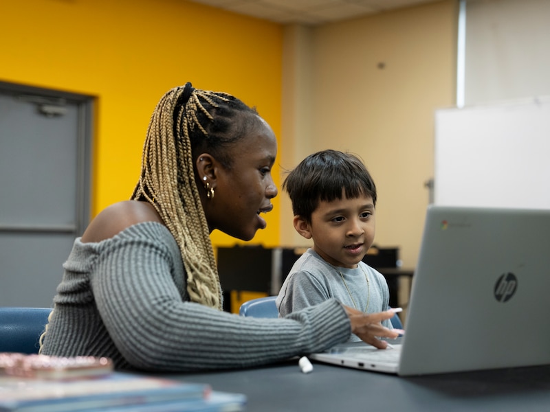 A child is sitting at a desk with an adult, and both of them are looking at the laptop on the desk.