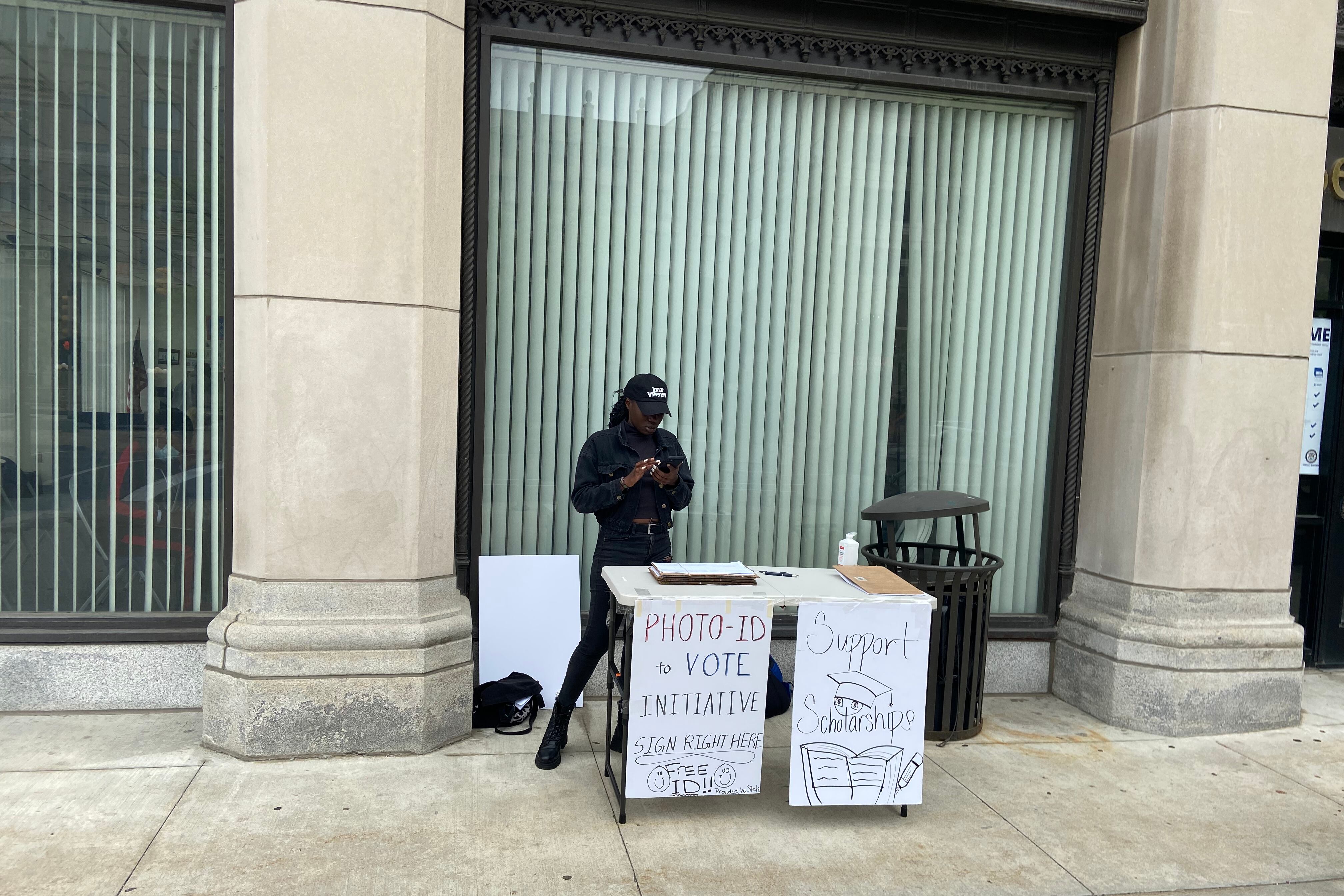 a person stands behind a table with hand written signs saying support petitions and free IDs