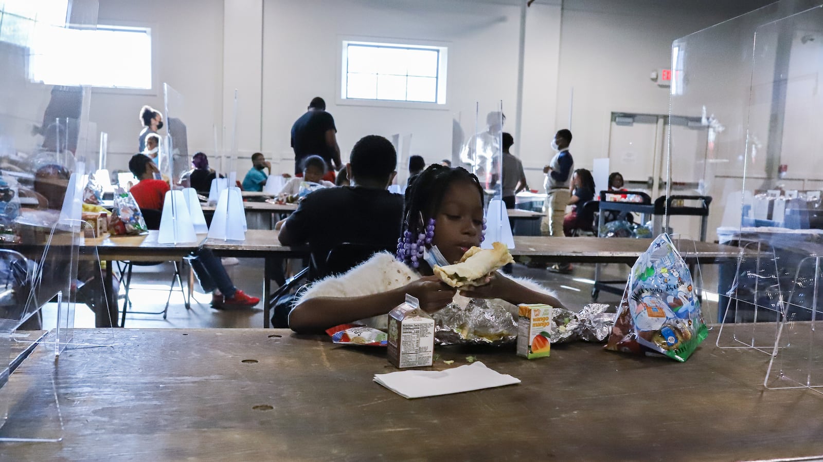 A young girl eats lunch while sitting in-between two large plastic barriers, as other students dine nearby.