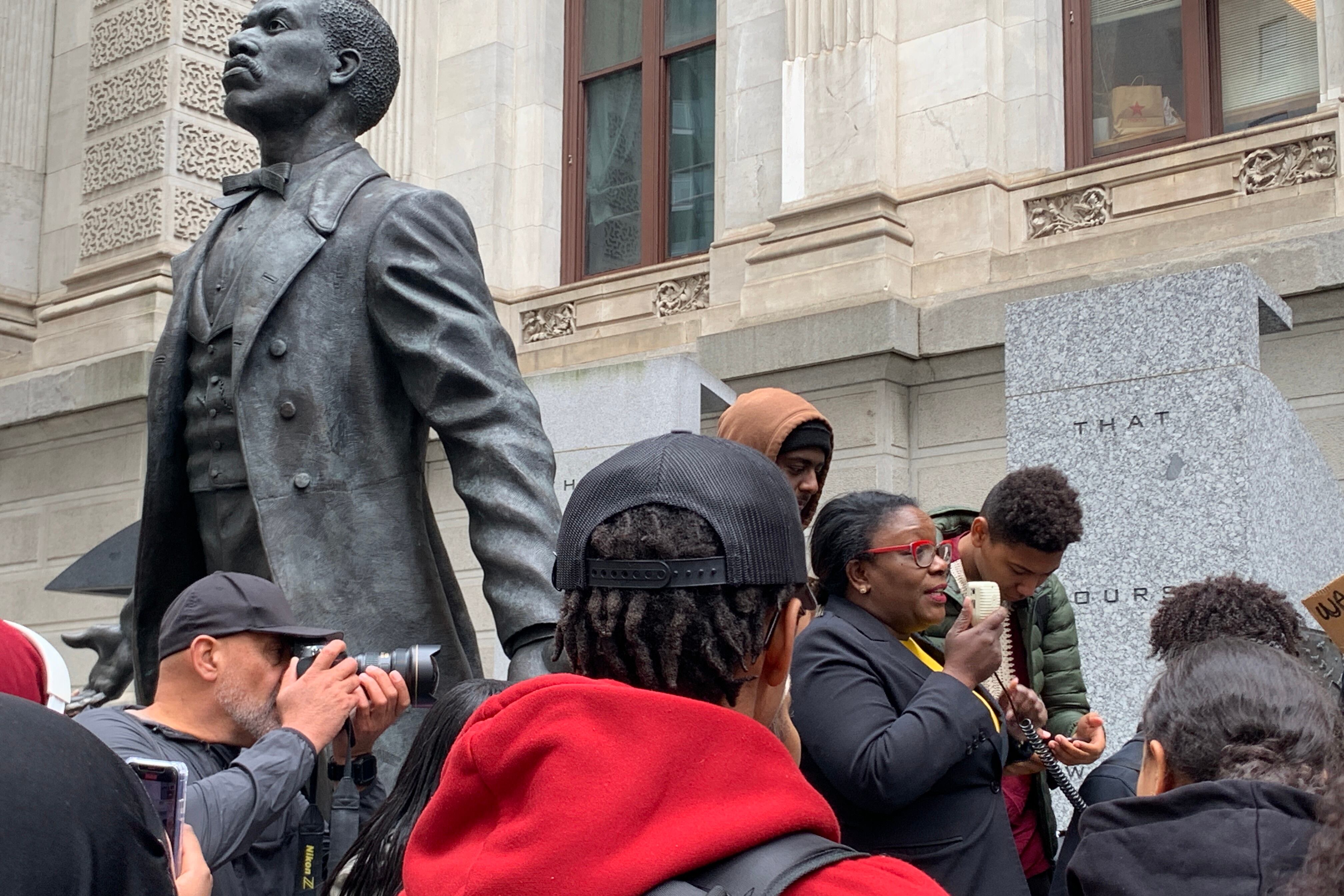 A woman wearing red glasses speaks into a PA system in front of a statue of a man.