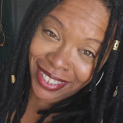 A photograph of a Black women smiling for a photo with long dark hair with two golden charms.