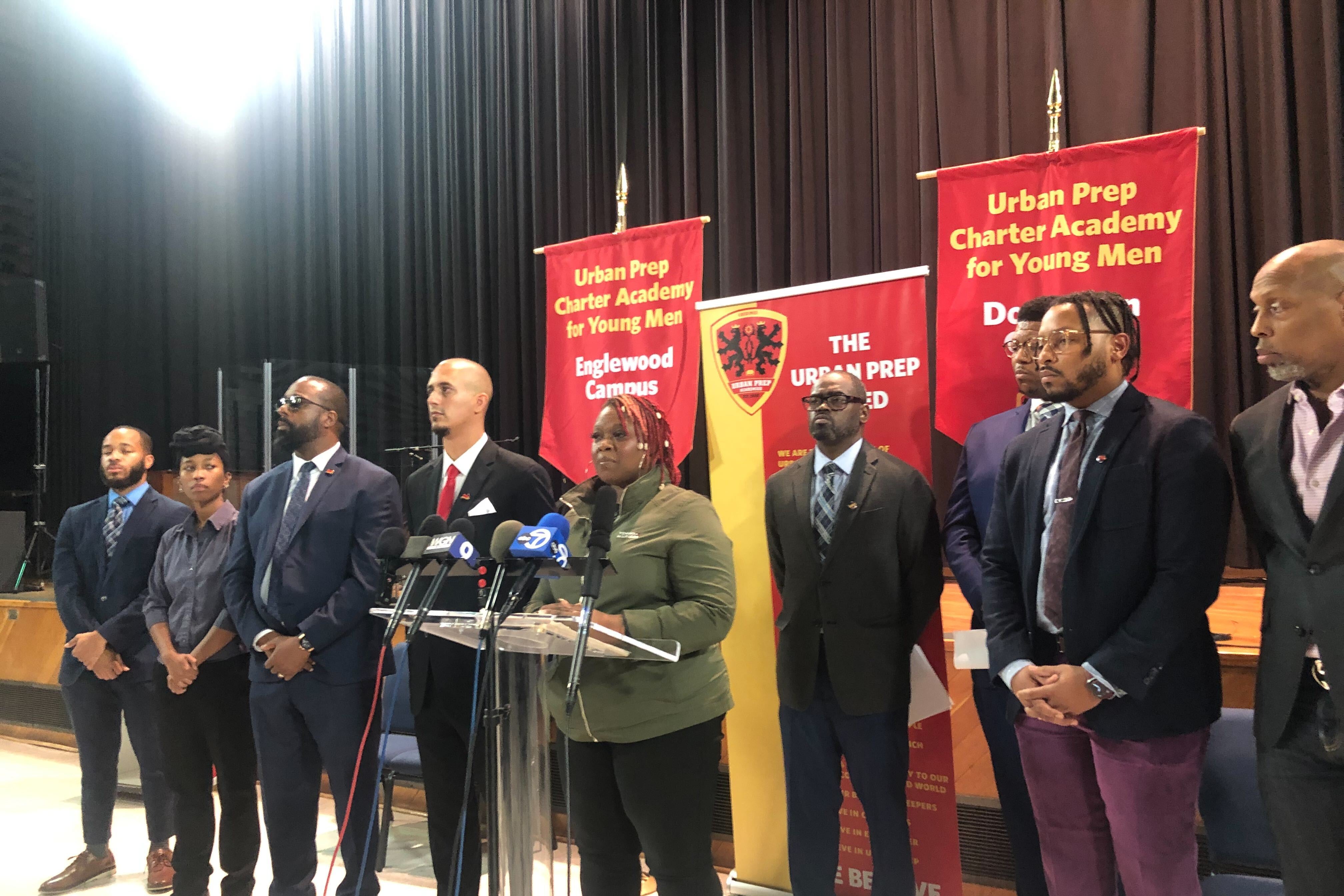 Men dressed in suits and a woman stand by a lectern in front of banners.