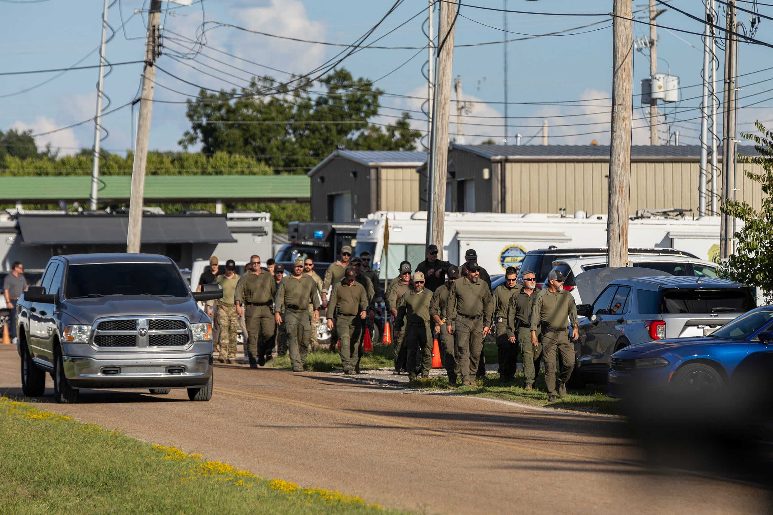 A photograph of a group of mostly white men wearing all green walking down a street with cars parked or driving on both sides of them.