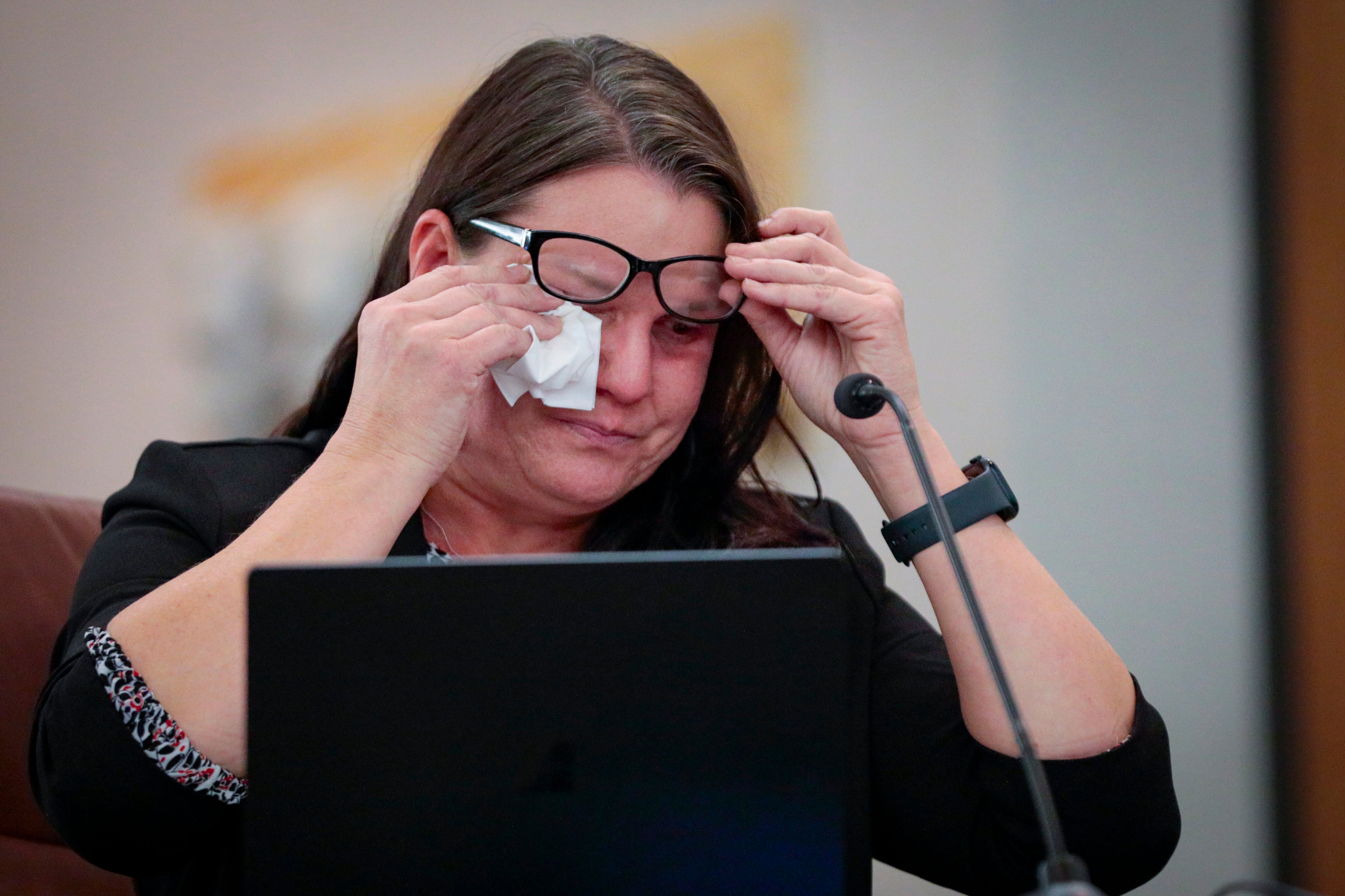 A woman with dark hair lifts her glasses to wipe her eyes with a tissue. There’s a laptop and mic in front of her.