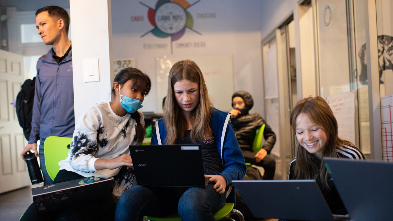 Three young teenage girls sit in chairs holding laptops. The girl on the left leans over to see the screen of the girl next to her, who seems to be concentrating. The girl on the right has a big smile. To the left of the image, an adult man in a button up shirt and short hair leans on a column.