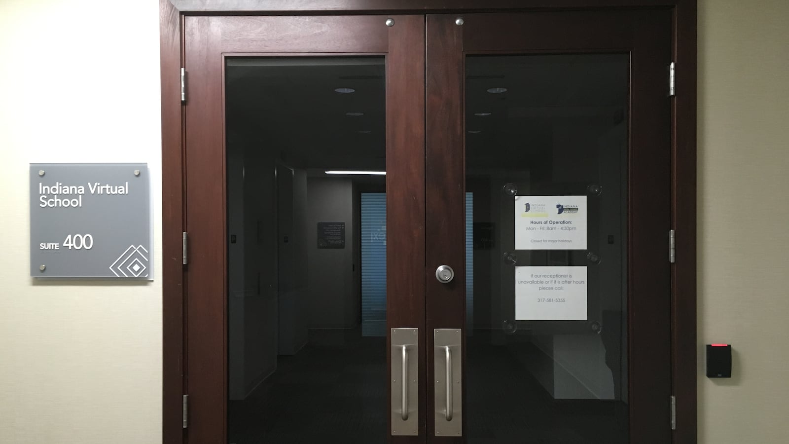 A close up of closed office doors made of dark wood with a light wall. There are signs hanging on the wall and the glass of the doors.