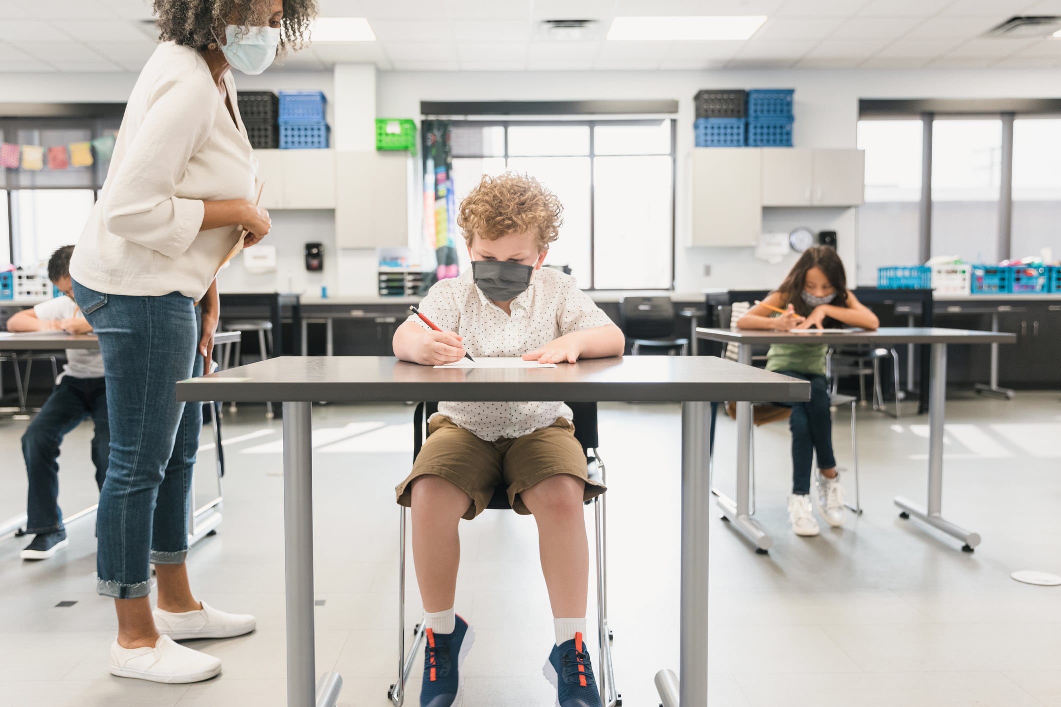 A young student wearing a mask sits in a classroom with a teacher standing nearby.