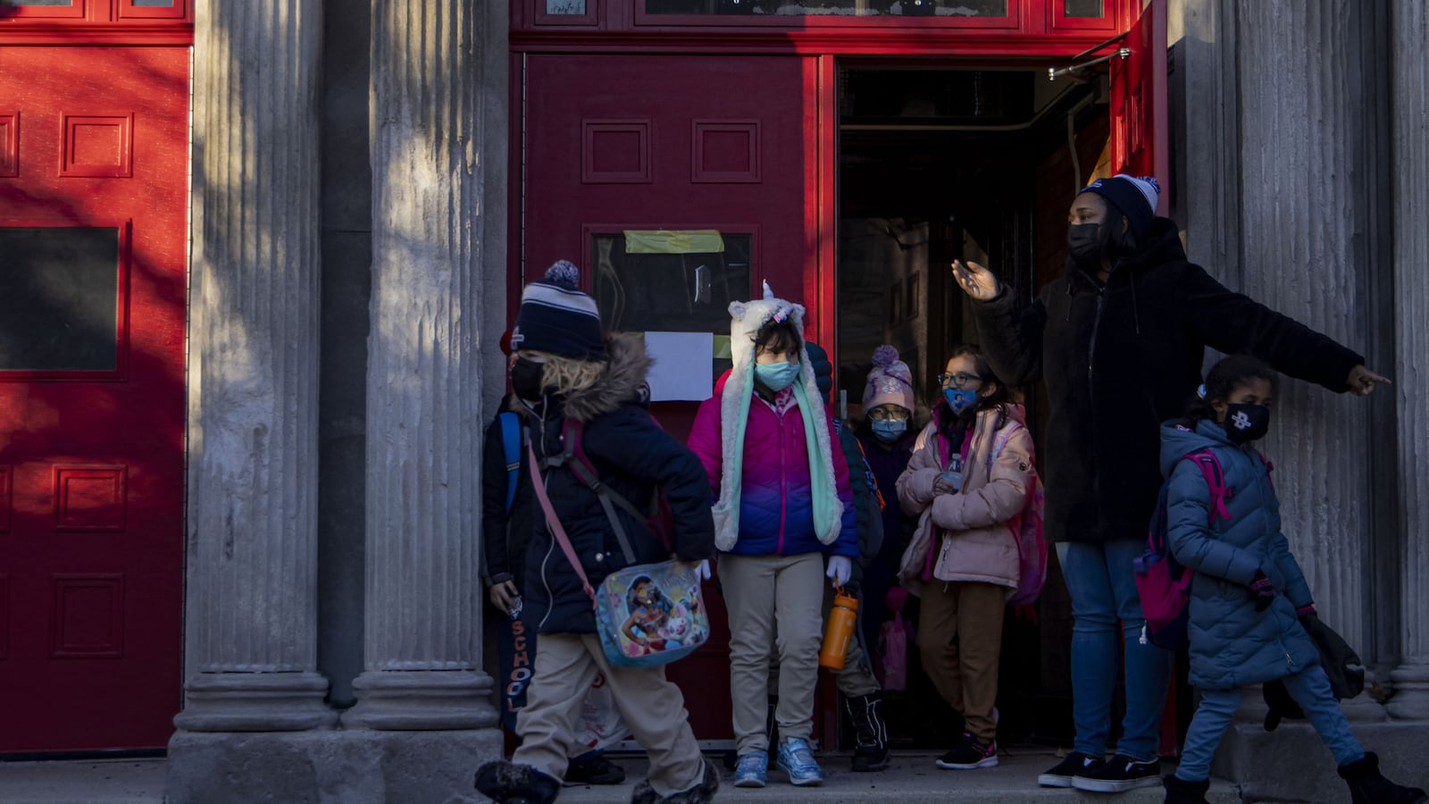 Students wearing winter clothes walk out of a red school door between gray stone columns.