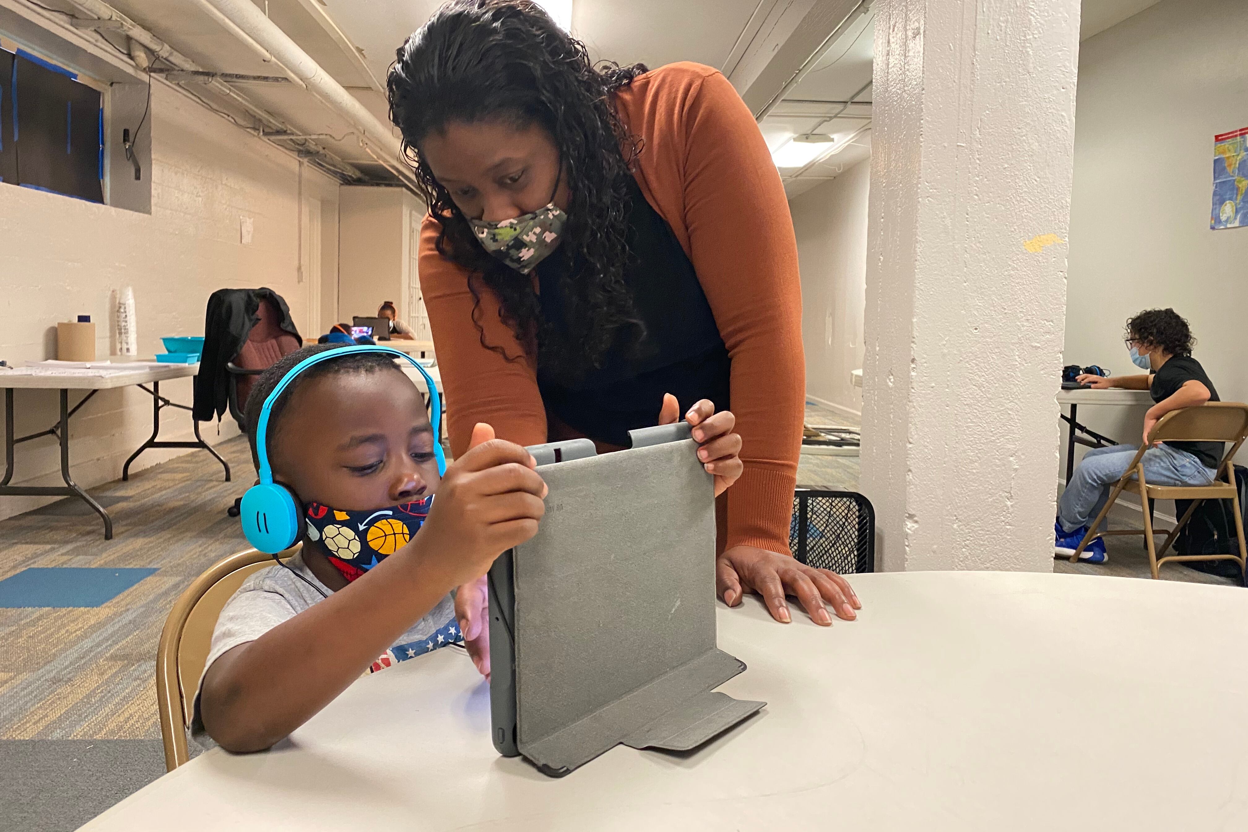 An adult e-learning specialist looks down on a student to help him log in to virtual class on his tablet.