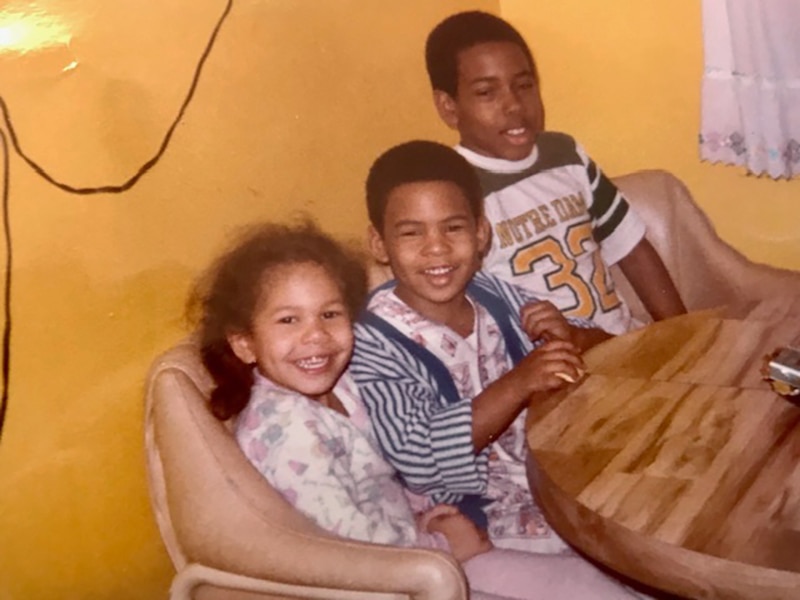 A photograph of an older photo of three young Black children smiling at the camera all sitting on a couch.