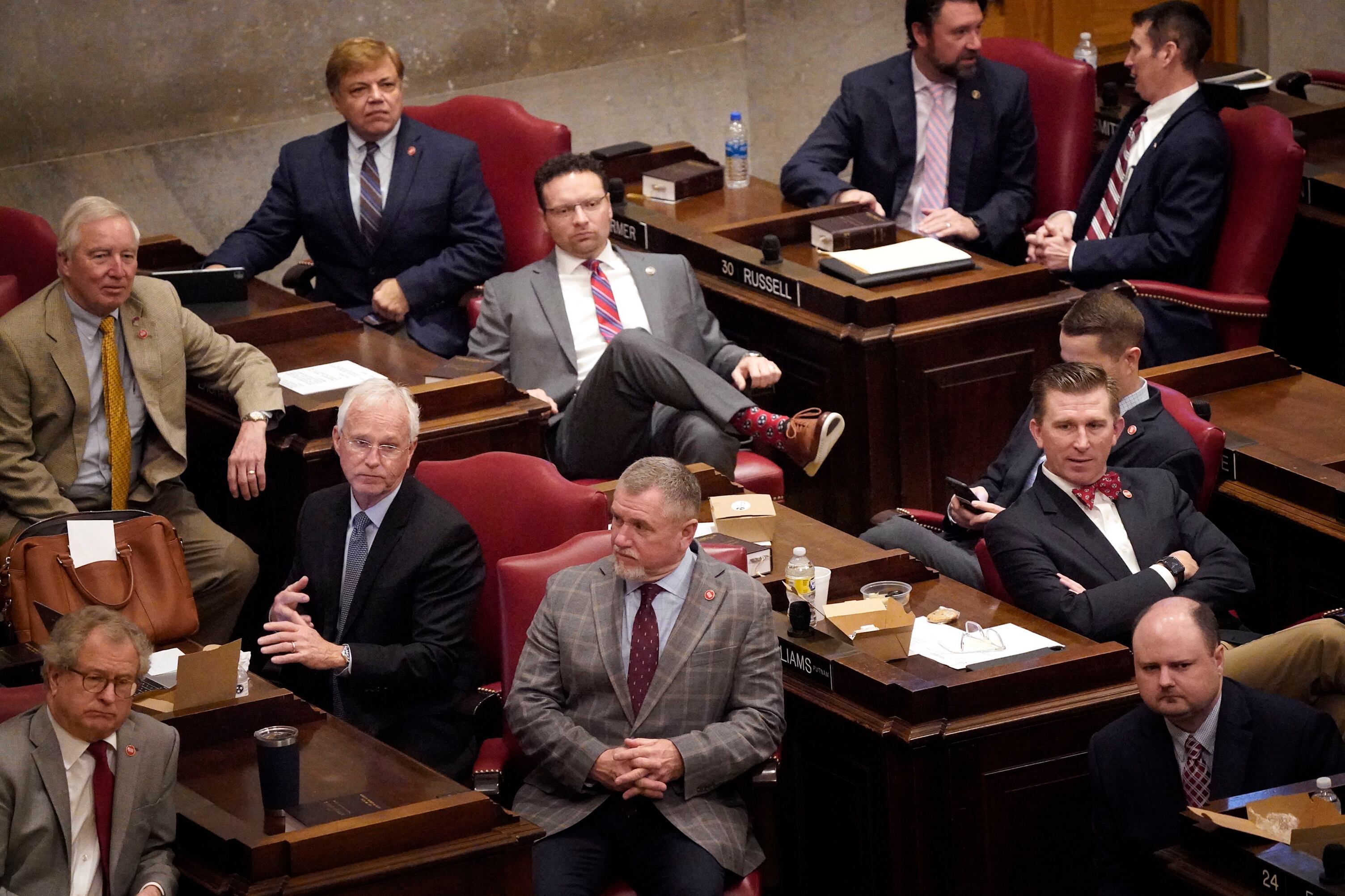 Men wearing business suits sit at desks in a large assembly hall.