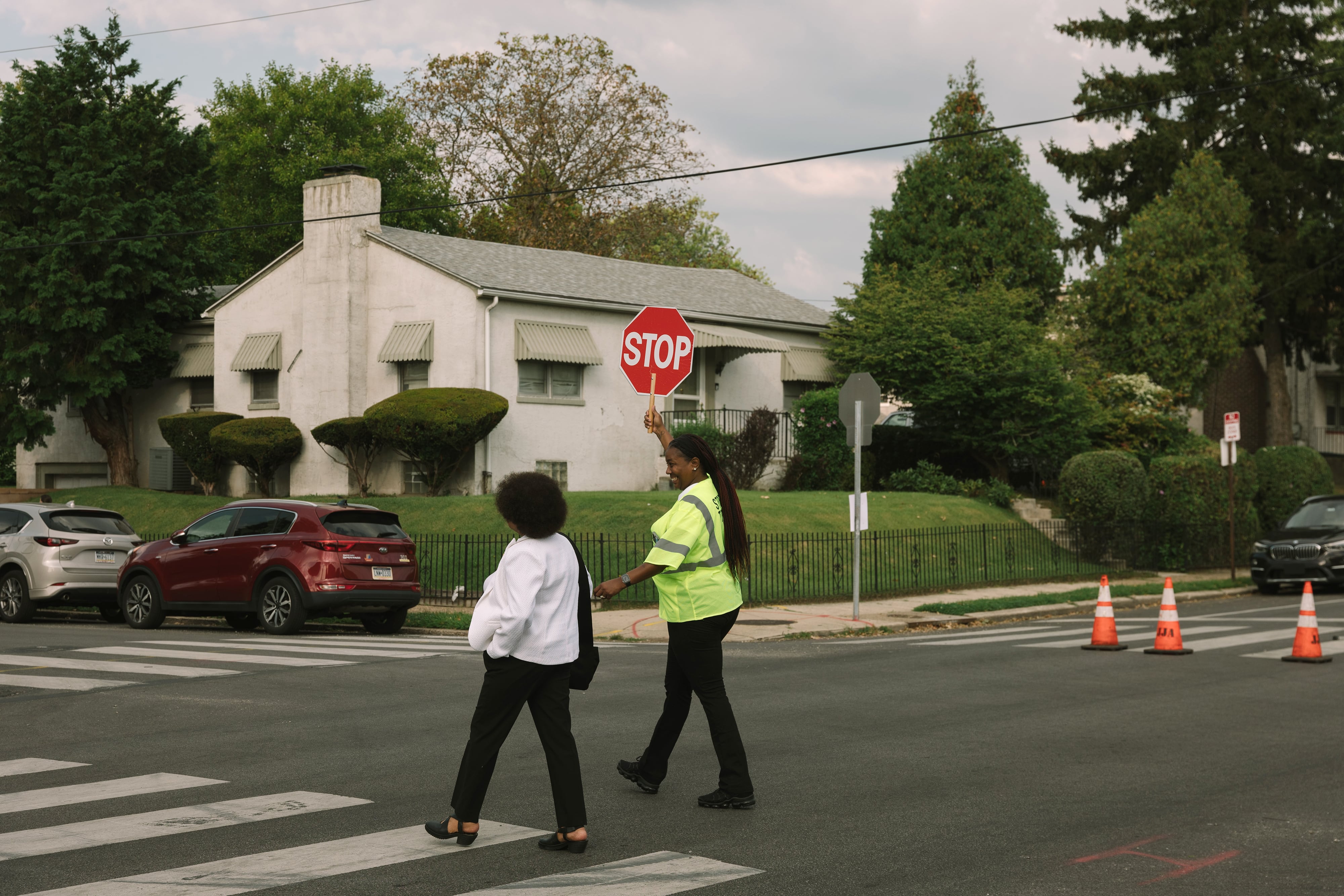 An adult crossing guard in a yellow vest and holding up a red stop sign helps a person wearing a white sweater cross a street.