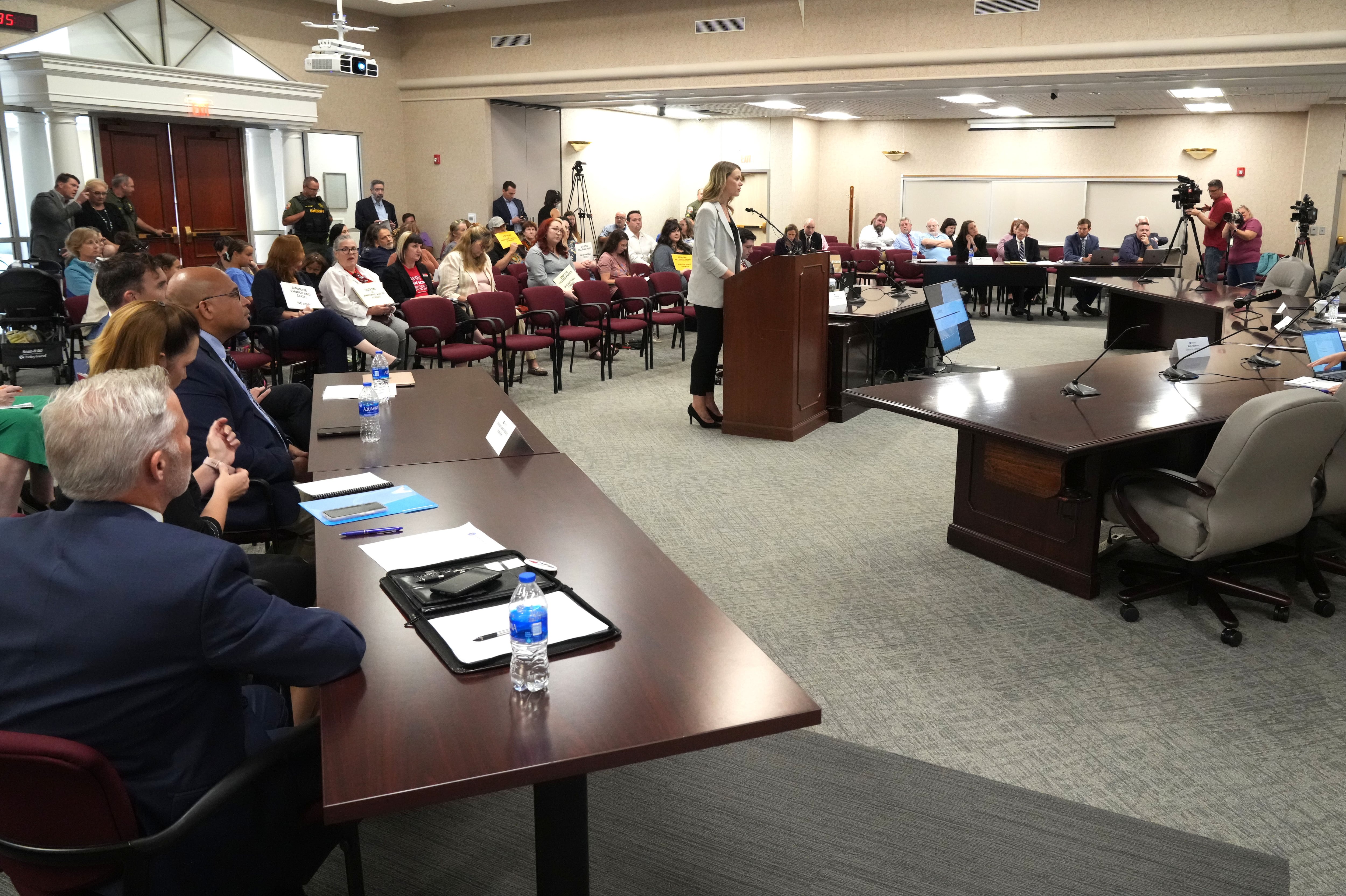 Woman speaks at a podium in large meeting filled with desks and people.