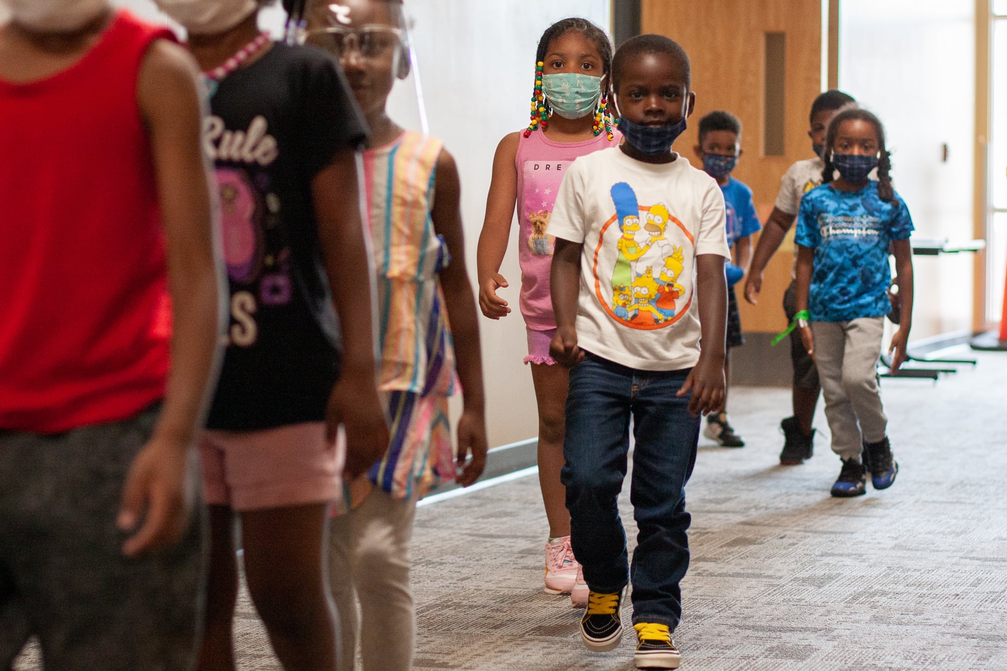 Eight kids, all wearing masks or face shields, walk in a jumbled line through a carpeted school hallway
