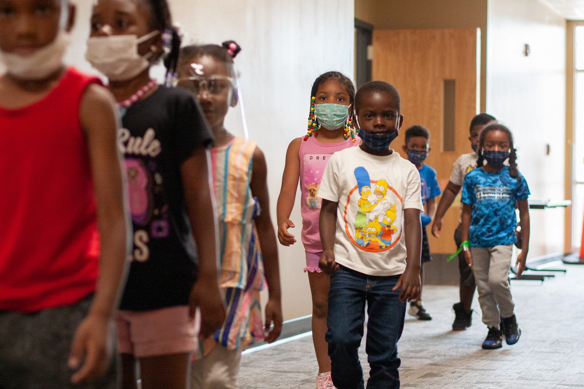 Eight kids, all wearing masks or face shields, walk in a jumbled line through a carpeted school hallway