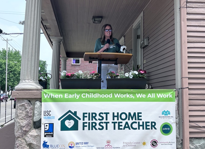 A white woman in a green t-shirt speaks from the porch of a house into a microphone outside.