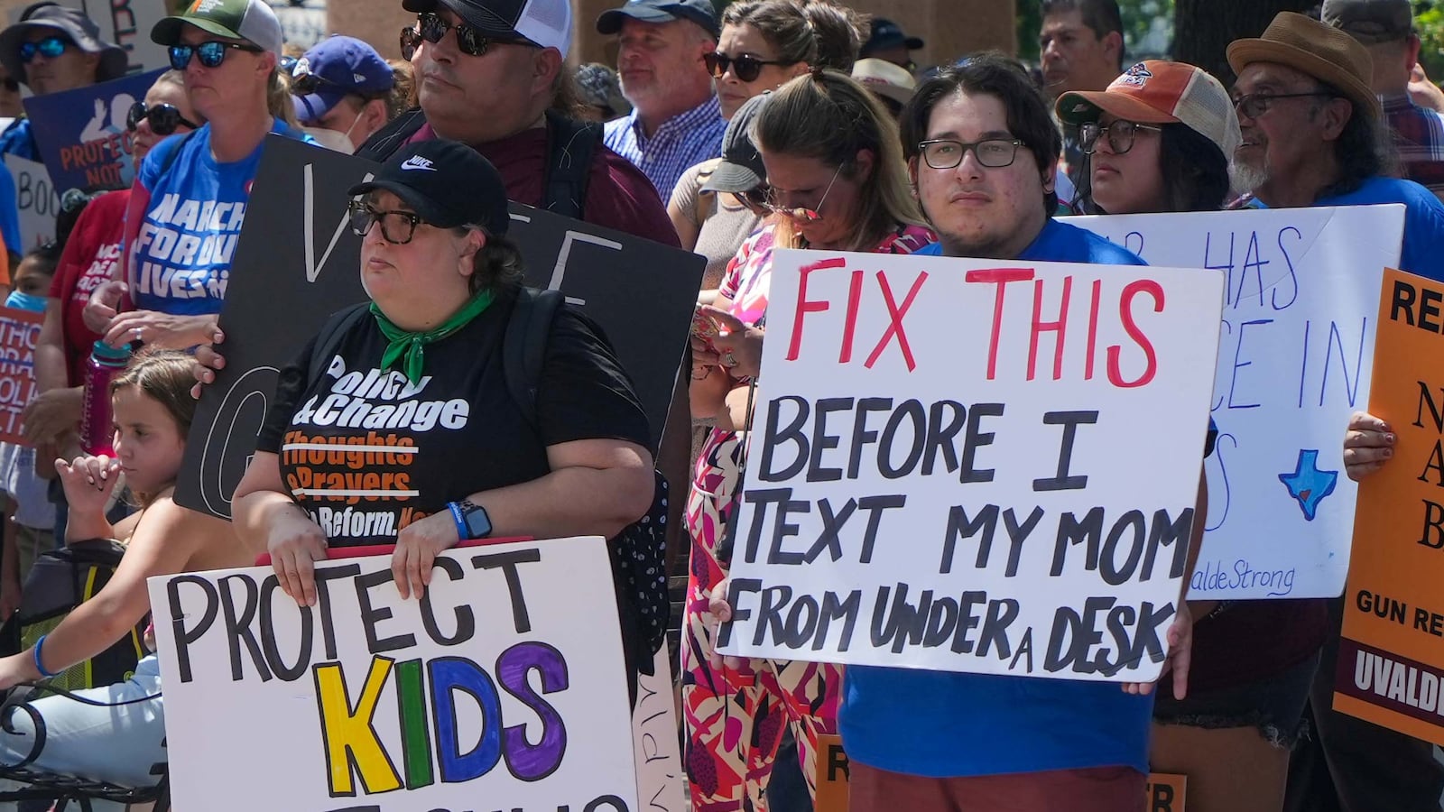 A group of people hold hand-made signs advocating for gun control. One sign says, "Protect Kids Not Guns." Another says "Fix This Before I text My Mom From Under Desk."