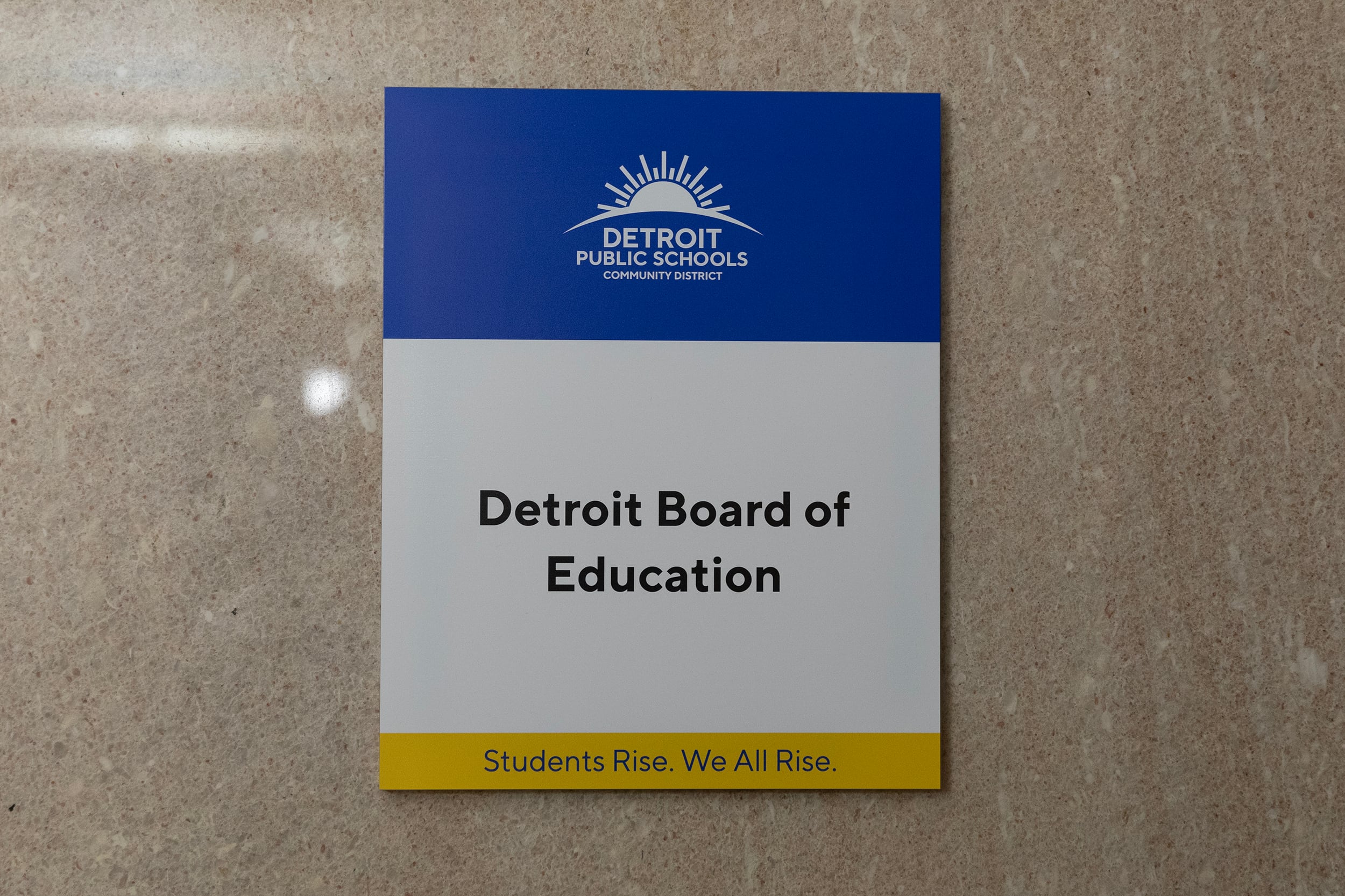 A small blue and white sign with black writing that reads "Detroit Board of Education" on the side of a marble wall.