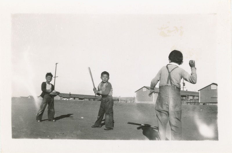 1 photographic print: b&w; Three young boys playing baseball outside of barracks at Amache concentration camp. There is a pitcher, batter, and catcher.