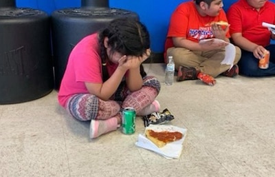 A young girl in a pink T-shirt sits on the floor of a community gym and holds her head in her hands, unable to eat the pizza or drink the soda in front of her.